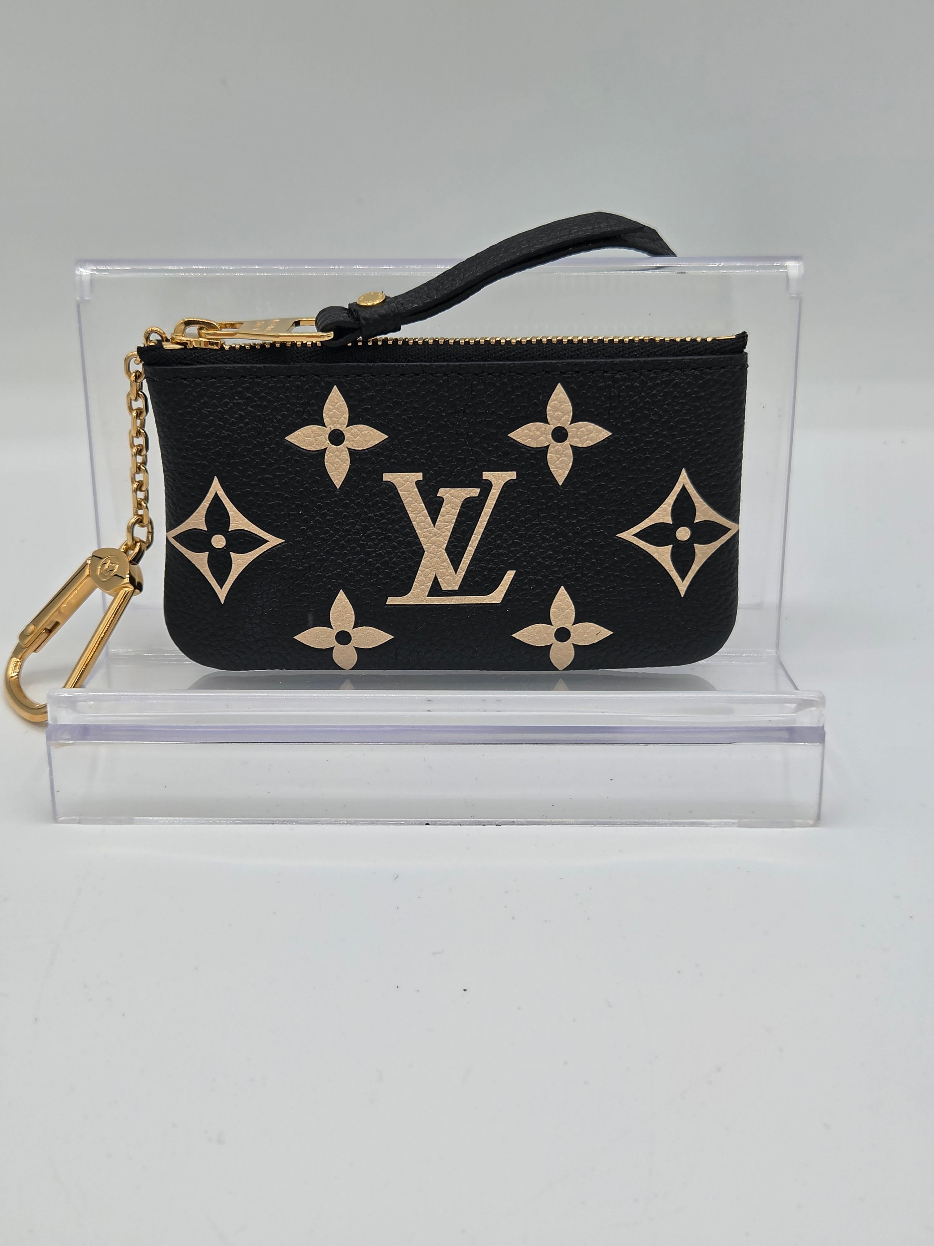 A Louis Vuitton black monogram wristlet pouch with gold-tone hardware, displayed inside a clear acrylic stand on a light gray background.