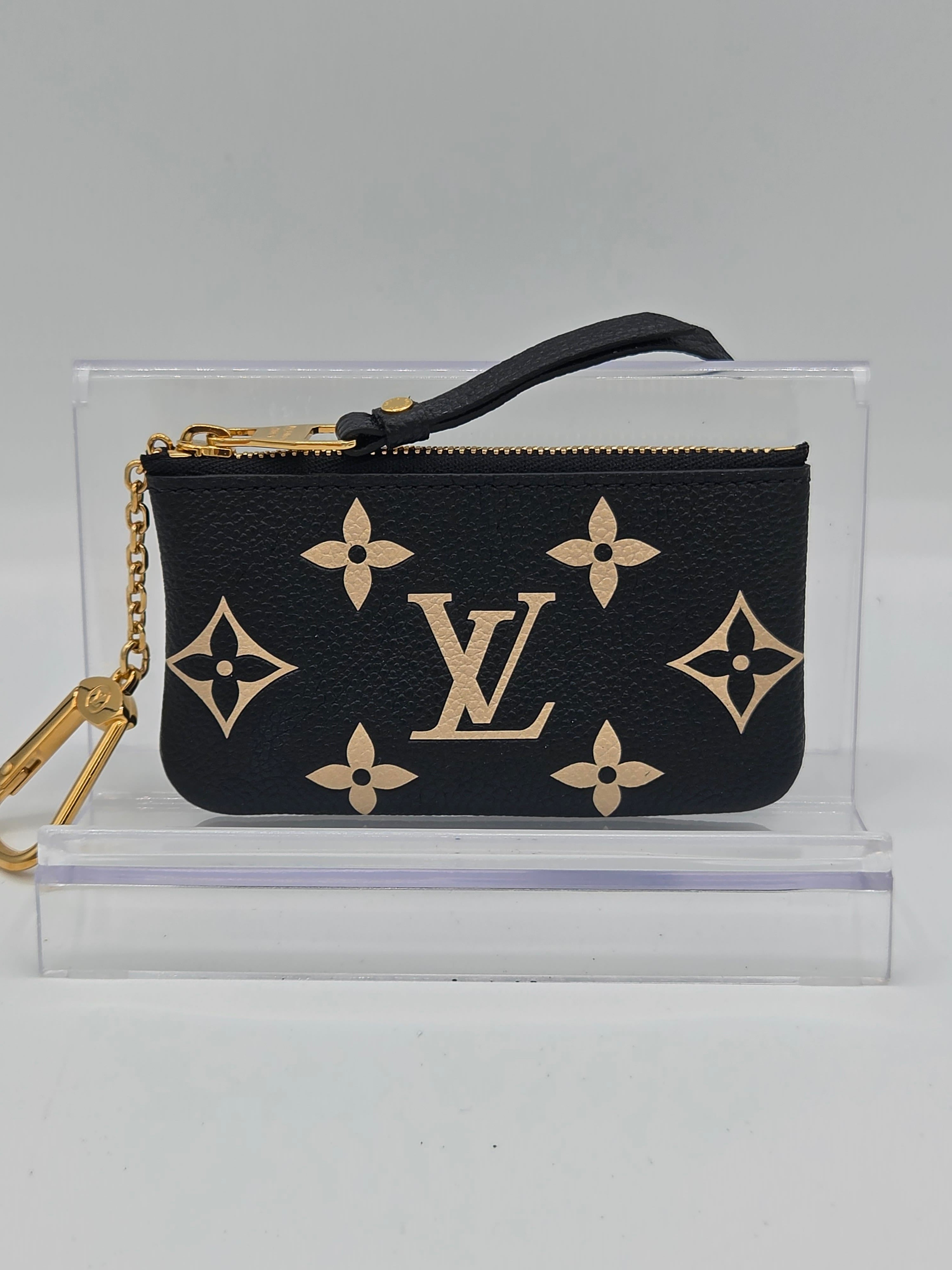 A Louis Vuitton monogram wristlet pouch in black coated canvas with gold-tone hardware, shown inside a clear acrylic display case against a light gray background.