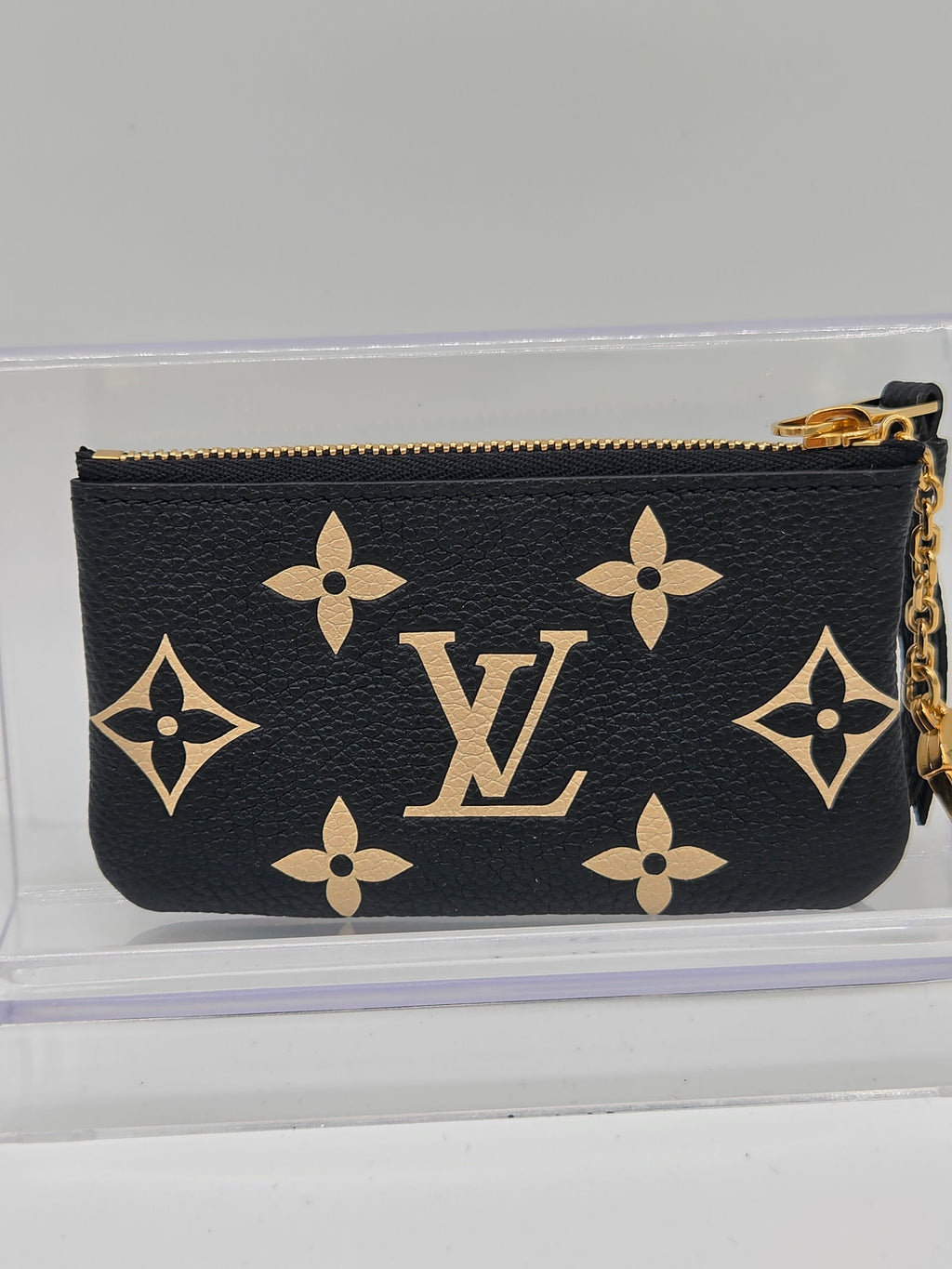 A Louis Vuitton monogram canvas coin pouch in black with gold-tone hardware and a gold chain strap, displayed inside a clear acrylic case.