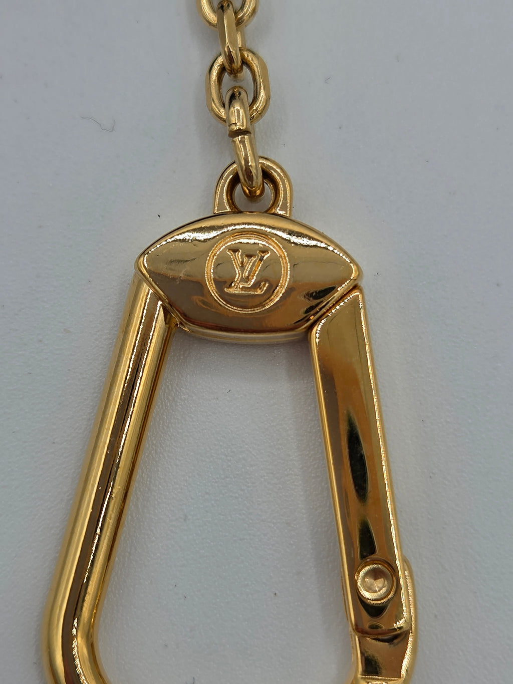 A close-up of a gold-tone metal carabiner-style keychain or bag charm with a visible Louis Vuitton monogram logo on the top plate, attached to a chain against a plain light background.