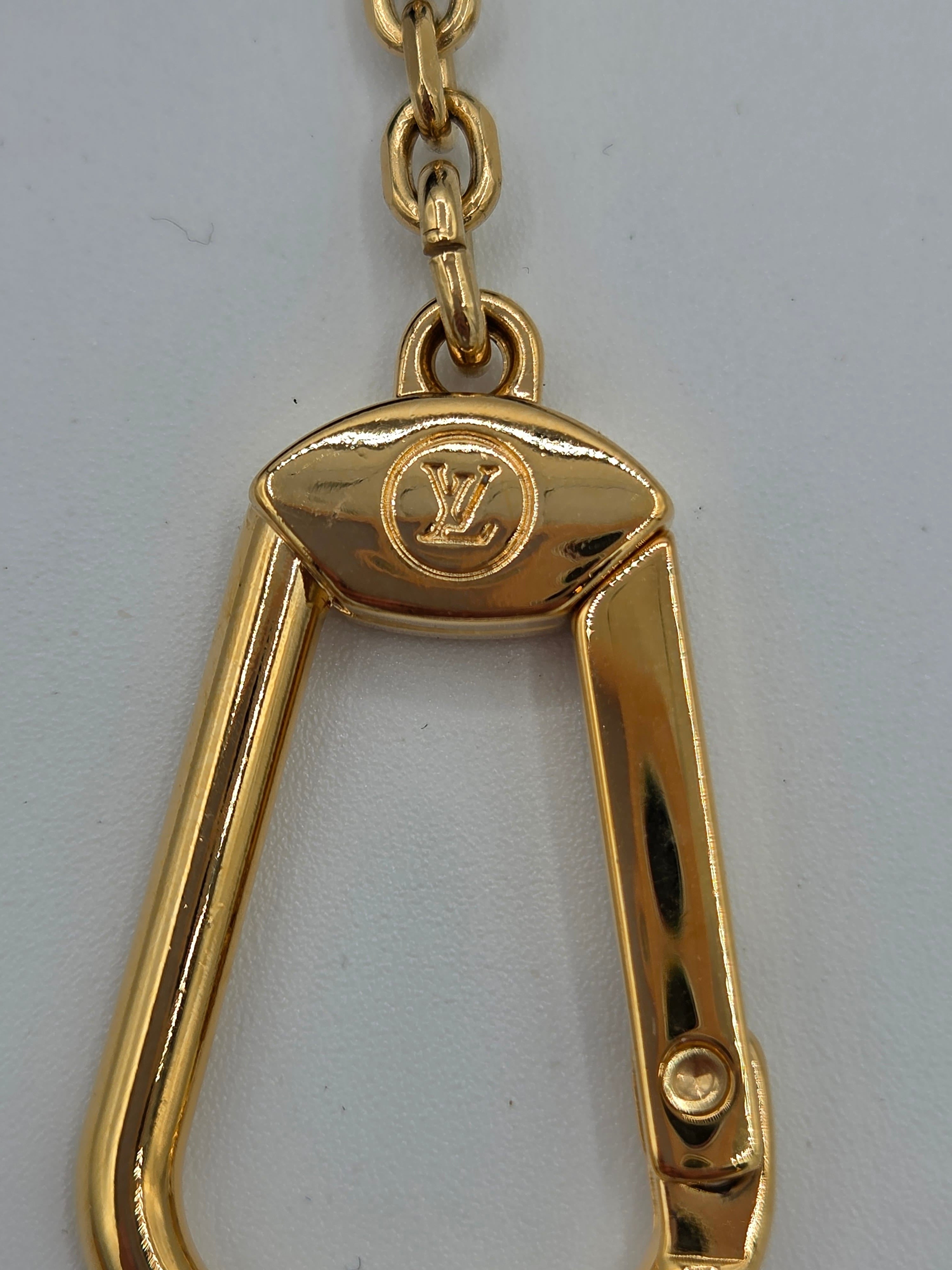 A close-up of a gold-tone metal carabiner-style keychain or bag charm with a visible Louis Vuitton monogram logo on the top plate, attached to a chain against a plain light background.