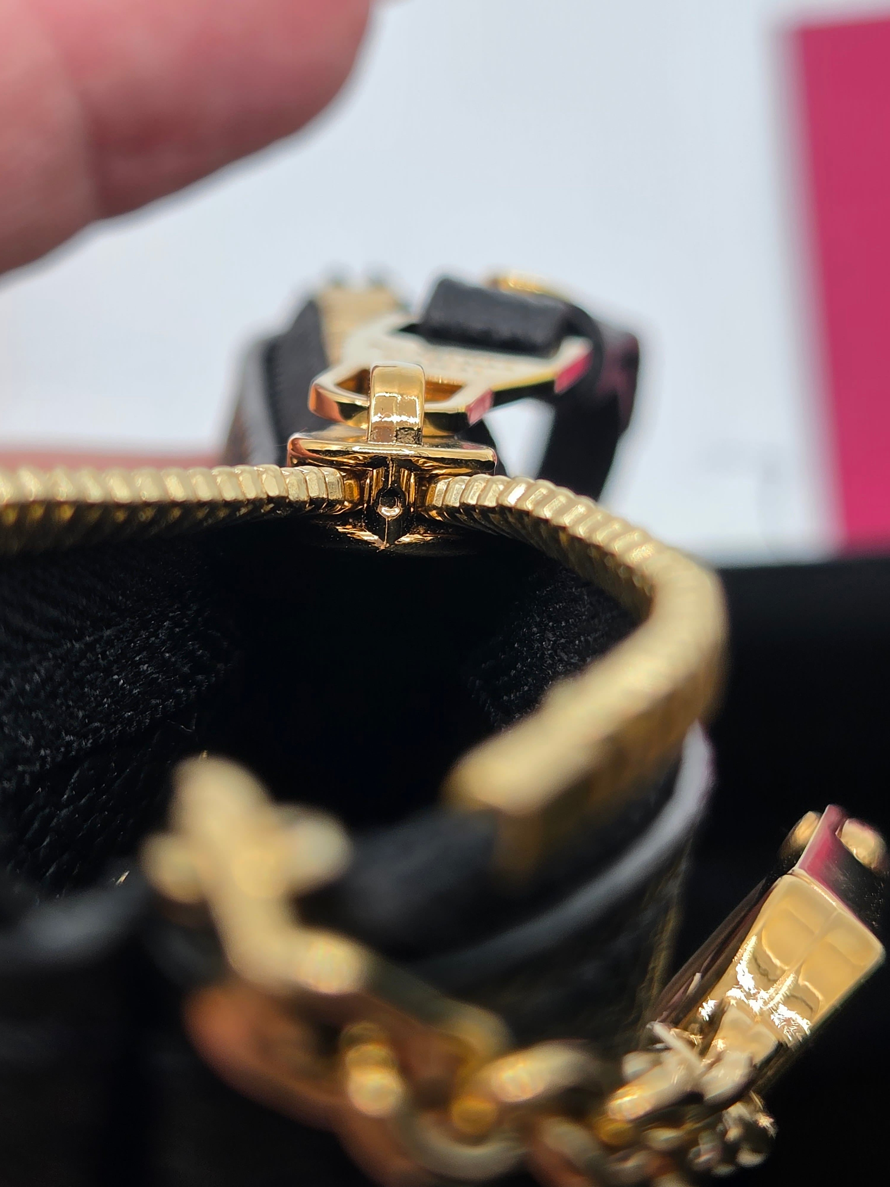 Close-up detail of a black handbag with gold-tone hardware, showing a textured zipper, clasp, and chain strap.