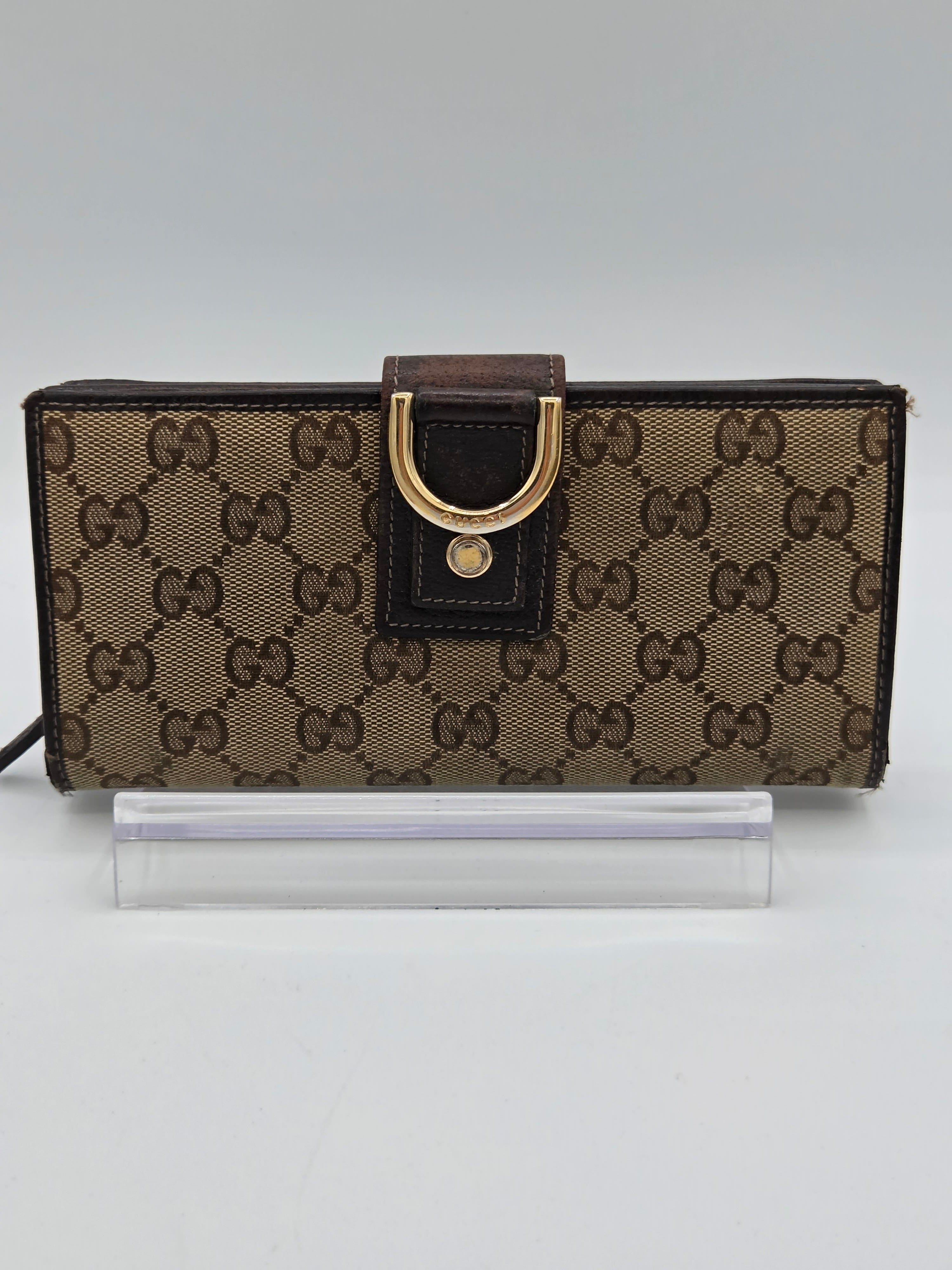 A Gucci monogram canvas wallet with a brown leather strap and gold-tone D-ring buckle, shown standing next to a clear acrylic block on a white background.