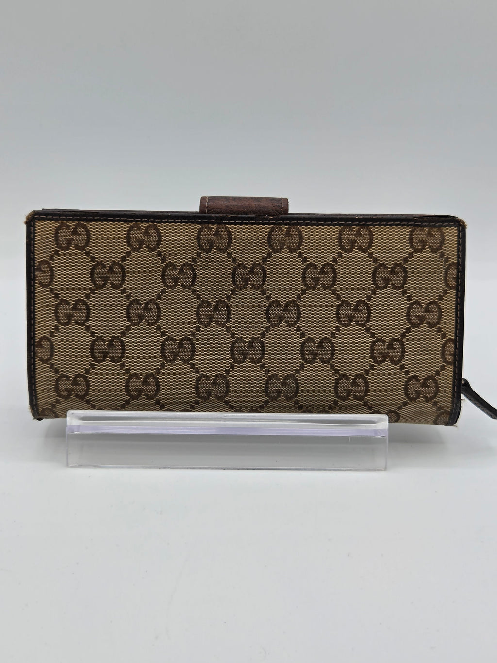 A Gucci monogram canvas wallet or phone case in brown and beige, shown standing vertically next to a clear plastic protective sleeve on a plain light background.