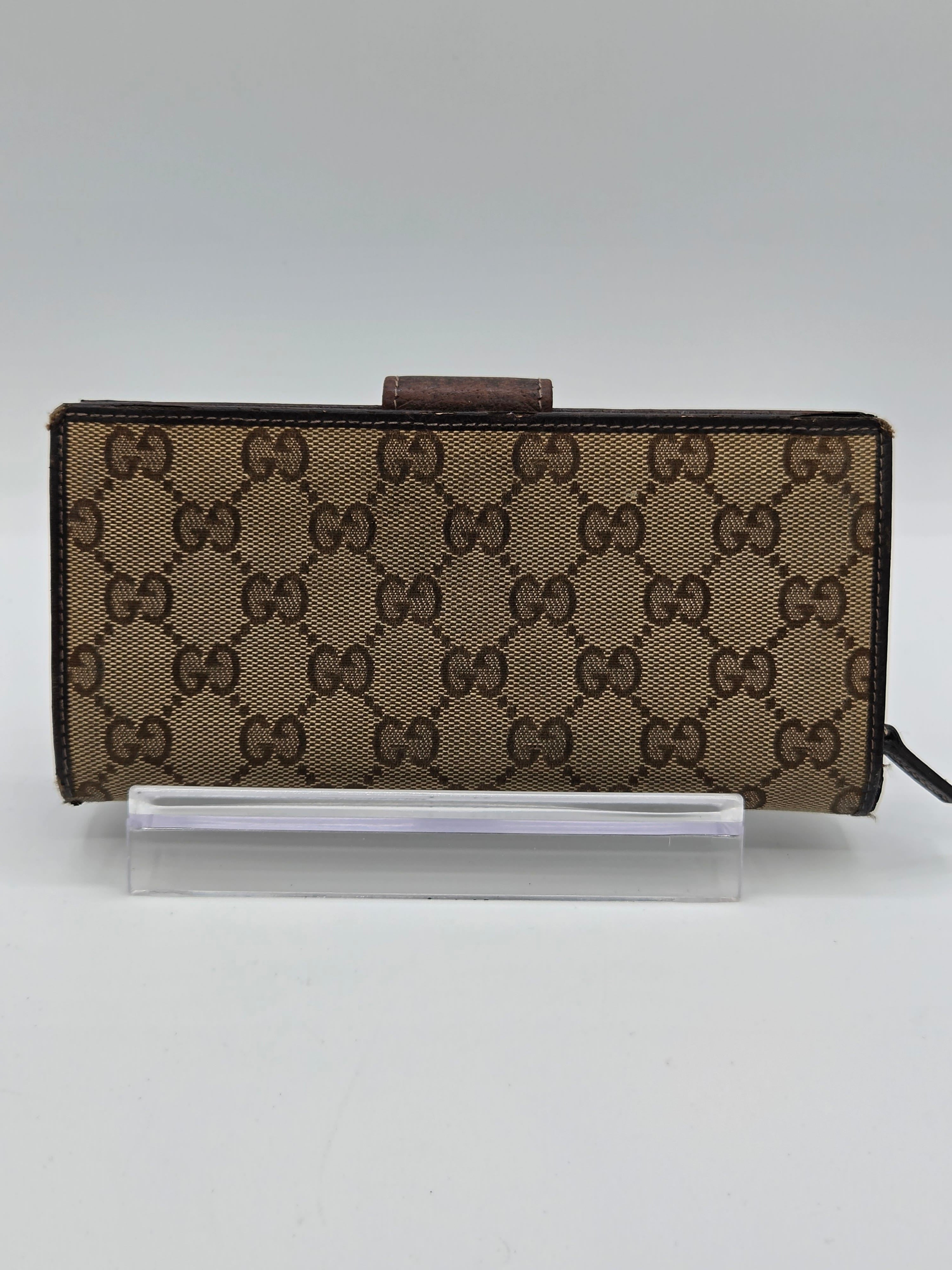 A Gucci monogram canvas wallet or phone case in brown and beige, shown standing vertically next to a clear plastic protective sleeve on a plain light background.