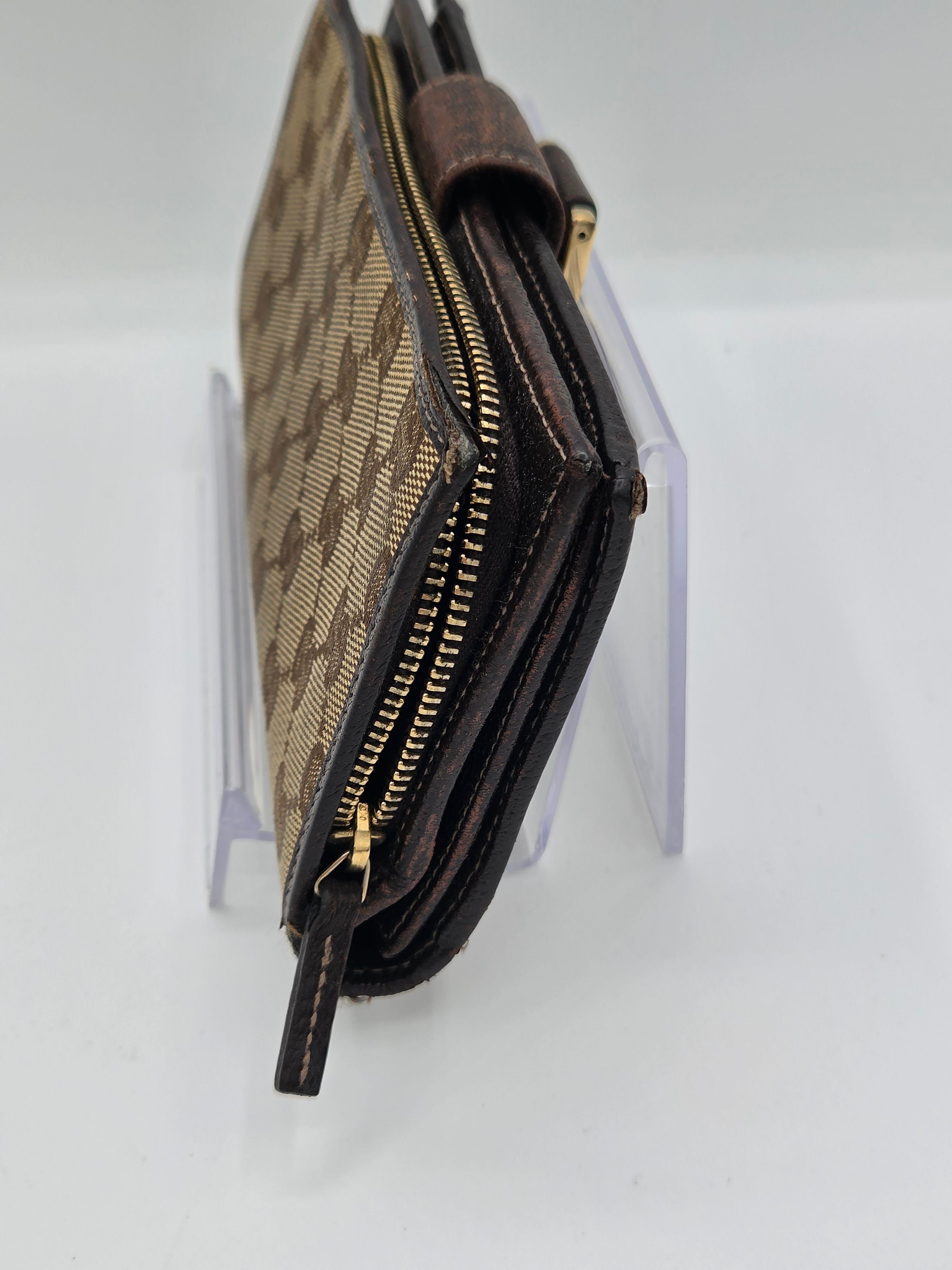 A close-up of a brown and beige patterned zip-around wallet or card holder with dark brown leather trim and gold-tone zipper hardware, displayed on a clear acrylic stand against a white background.