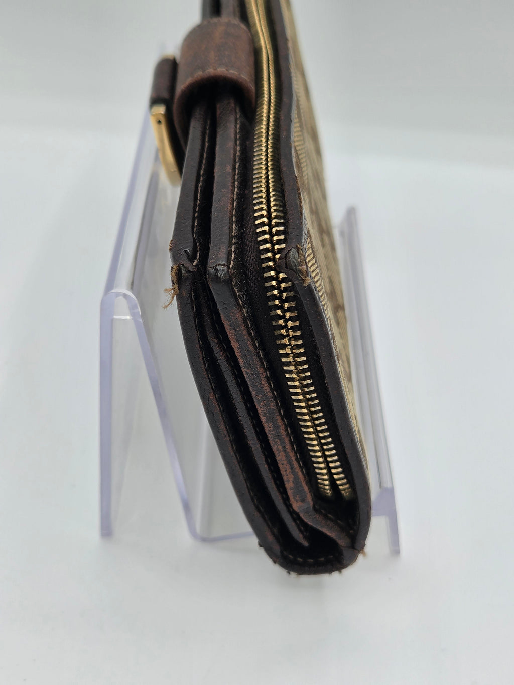 A close-up of a dark brown leather wallet or clutch with gold-tone zippers, resting on a clear acrylic display stand against a white background.