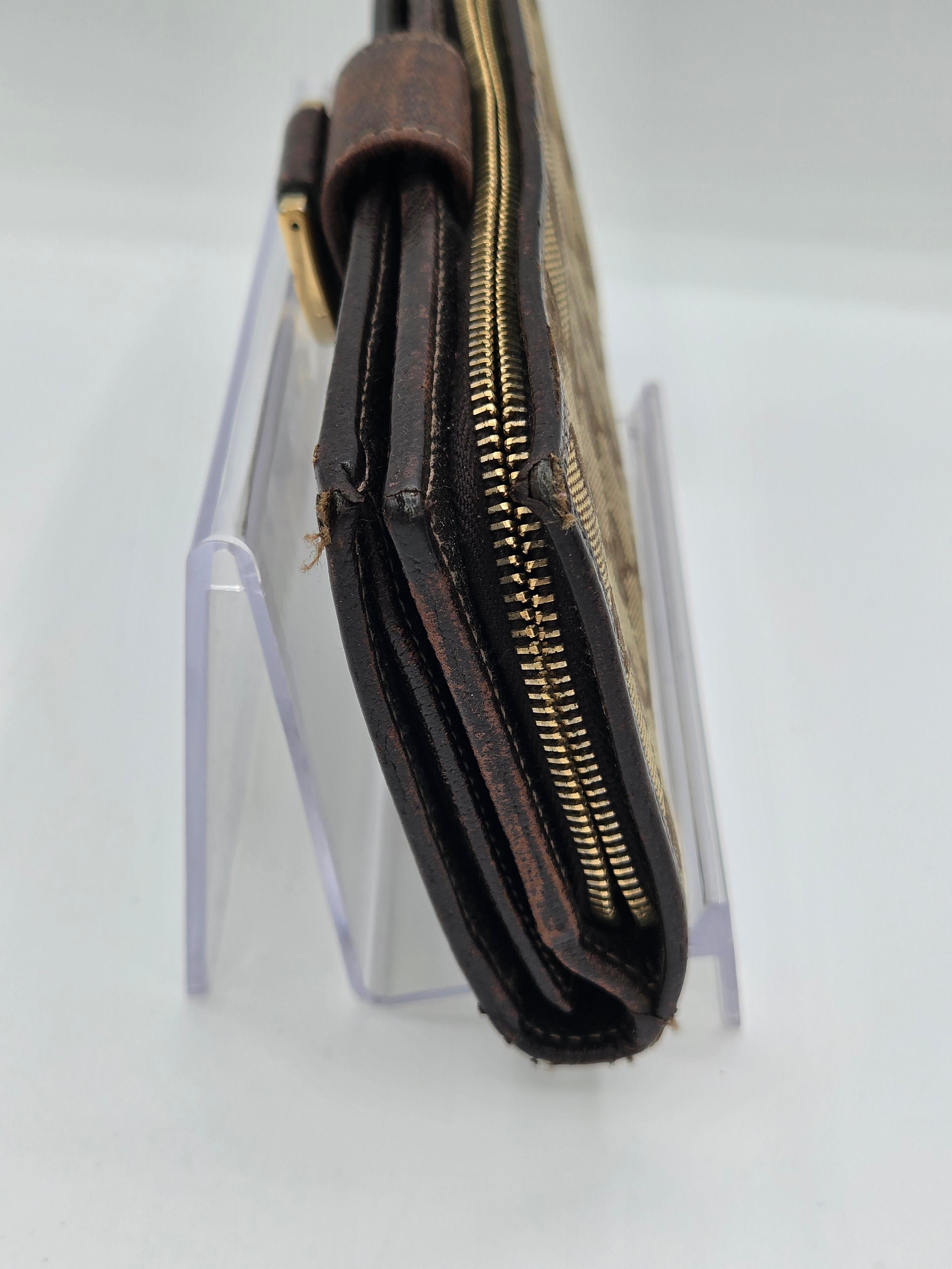 A close-up of a dark brown leather wallet or clutch with gold-tone zippers, resting on a clear acrylic display stand against a white background.