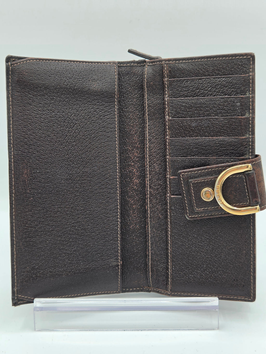 A dark brown textured leather wallet shown open, featuring multiple card slots, a zippered compartment, and a gold-tone metal D-ring closure on the right side, displayed on a clear acrylic stand against a light gray background.