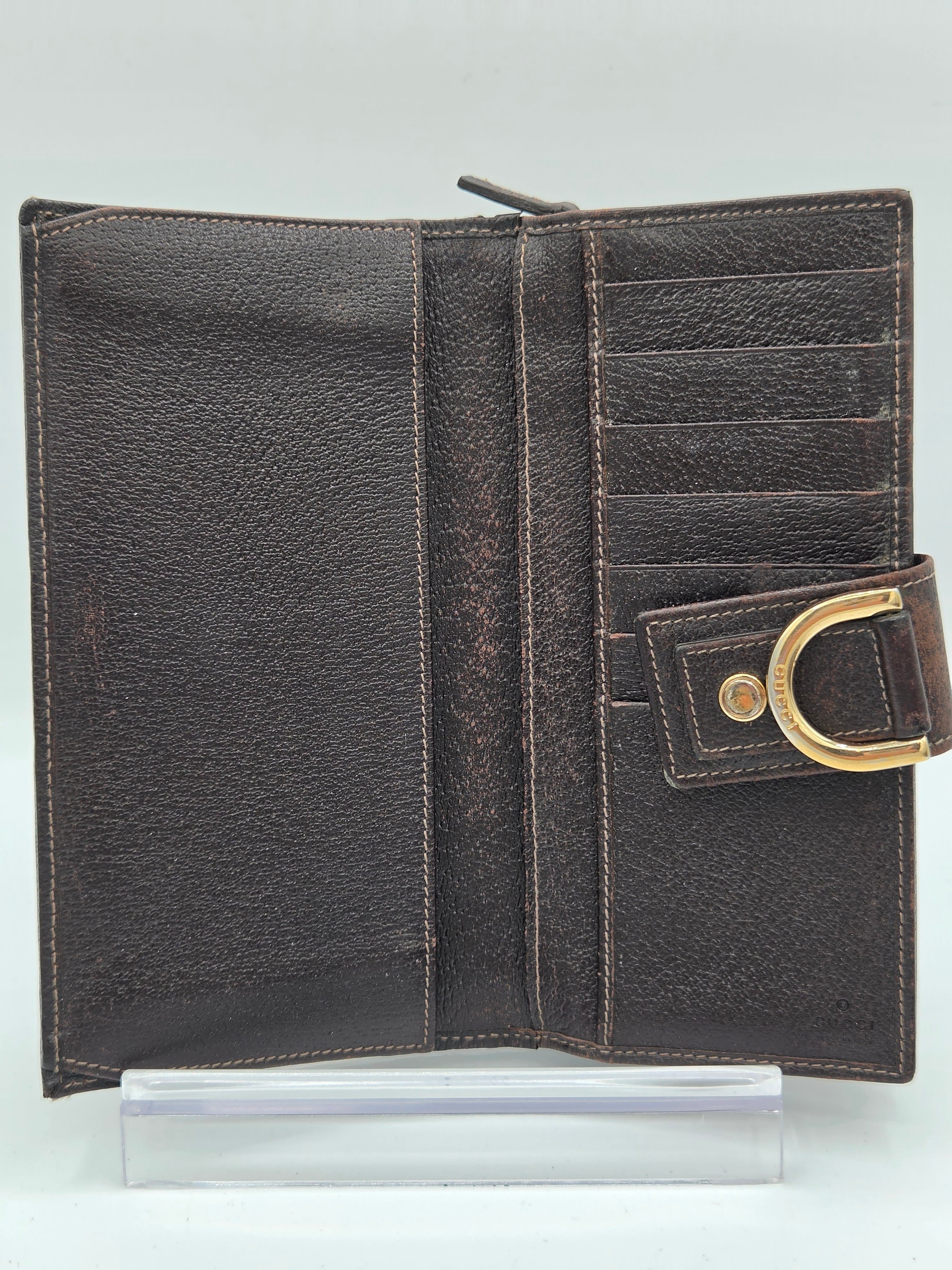 A dark brown textured leather wallet shown open, featuring multiple card slots, a zippered compartment, and a gold-tone metal D-ring closure on the right side, displayed on a clear acrylic stand against a light gray background.