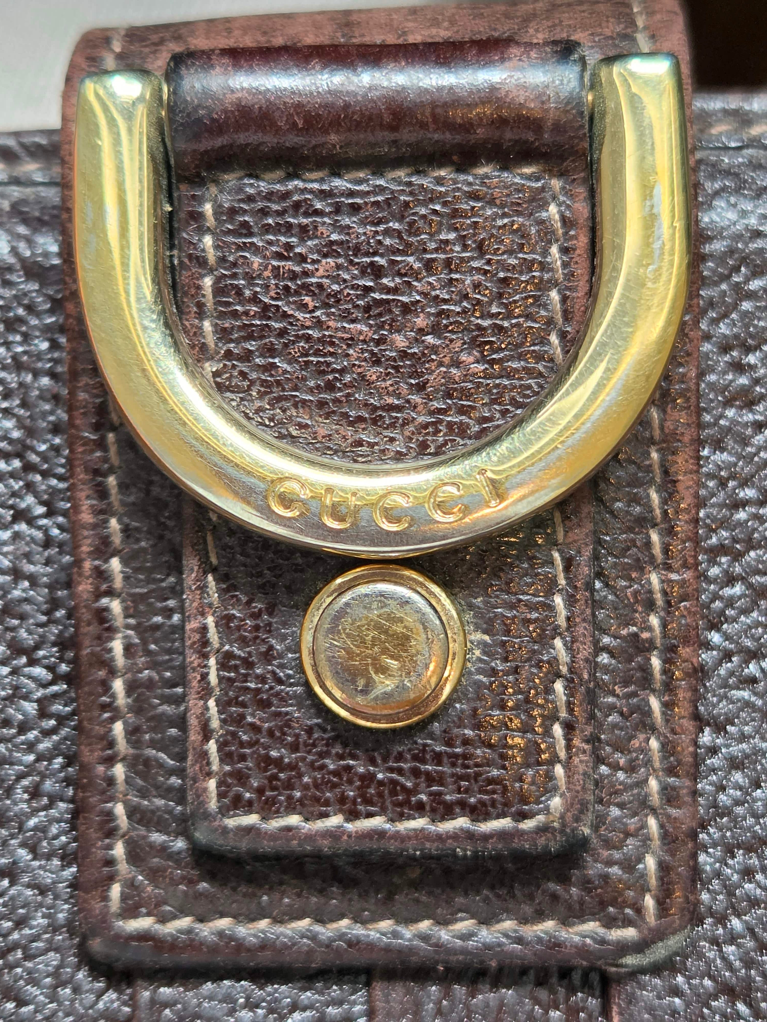 Close-up of a brown leather belt with a large gold-tone metal D-ring buckle and a round gold-tone rivet on a dark textured background.