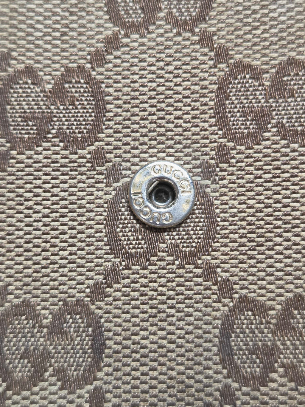 Close-up of a Gucci monogram canvas bag featuring the brand's signature GG pattern in beige and brown, with a silver-tone metal snap button in the center.