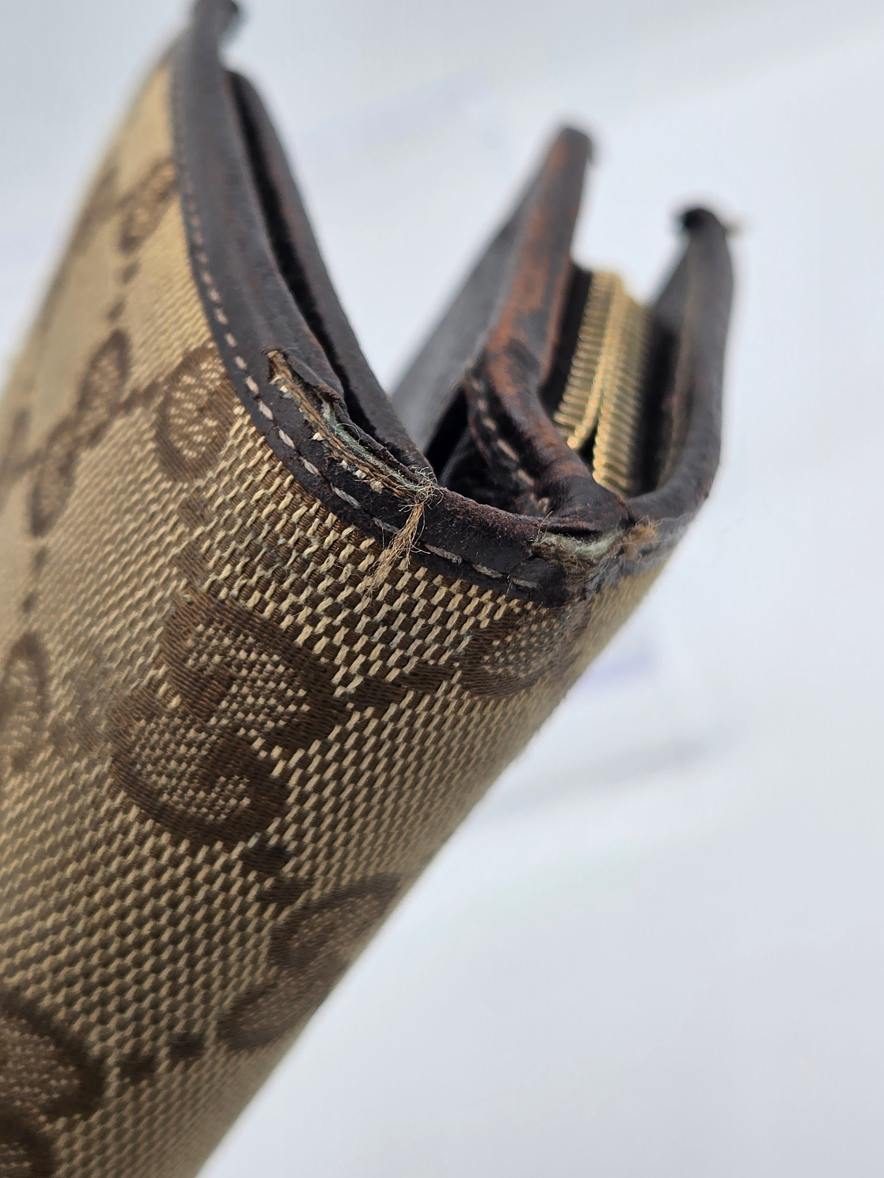 Close-up of a Gucci monogram canvas zippered wallet or card case, showing the beige and brown GG pattern, dark brown leather trim, and a gold-tone zipper pull against a white background.