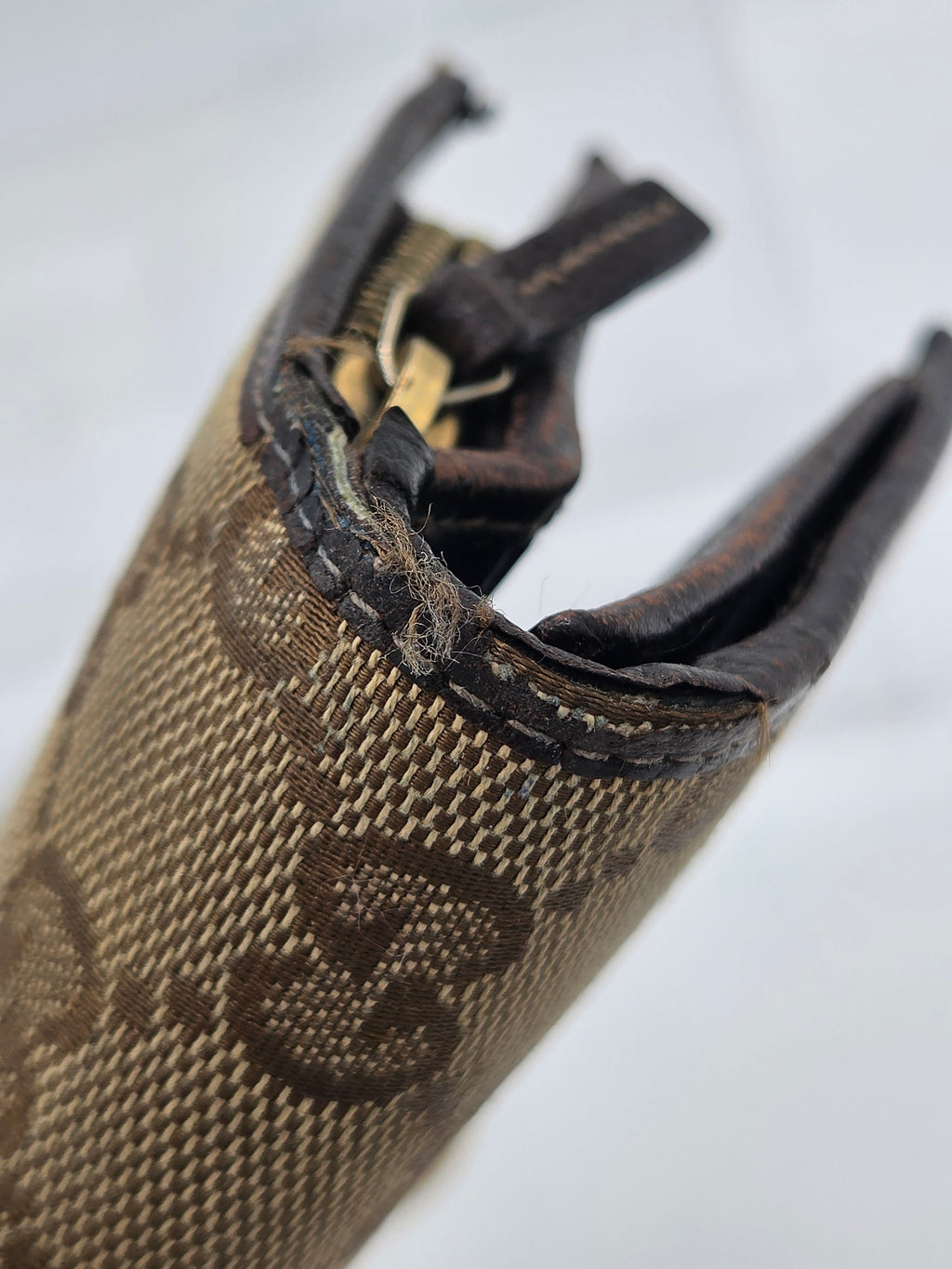 Close-up of a Gucci monogram canvas pouch showing a severely ripped and frayed zipper pull tab, with dark brown leather trim and the classic GG pattern.