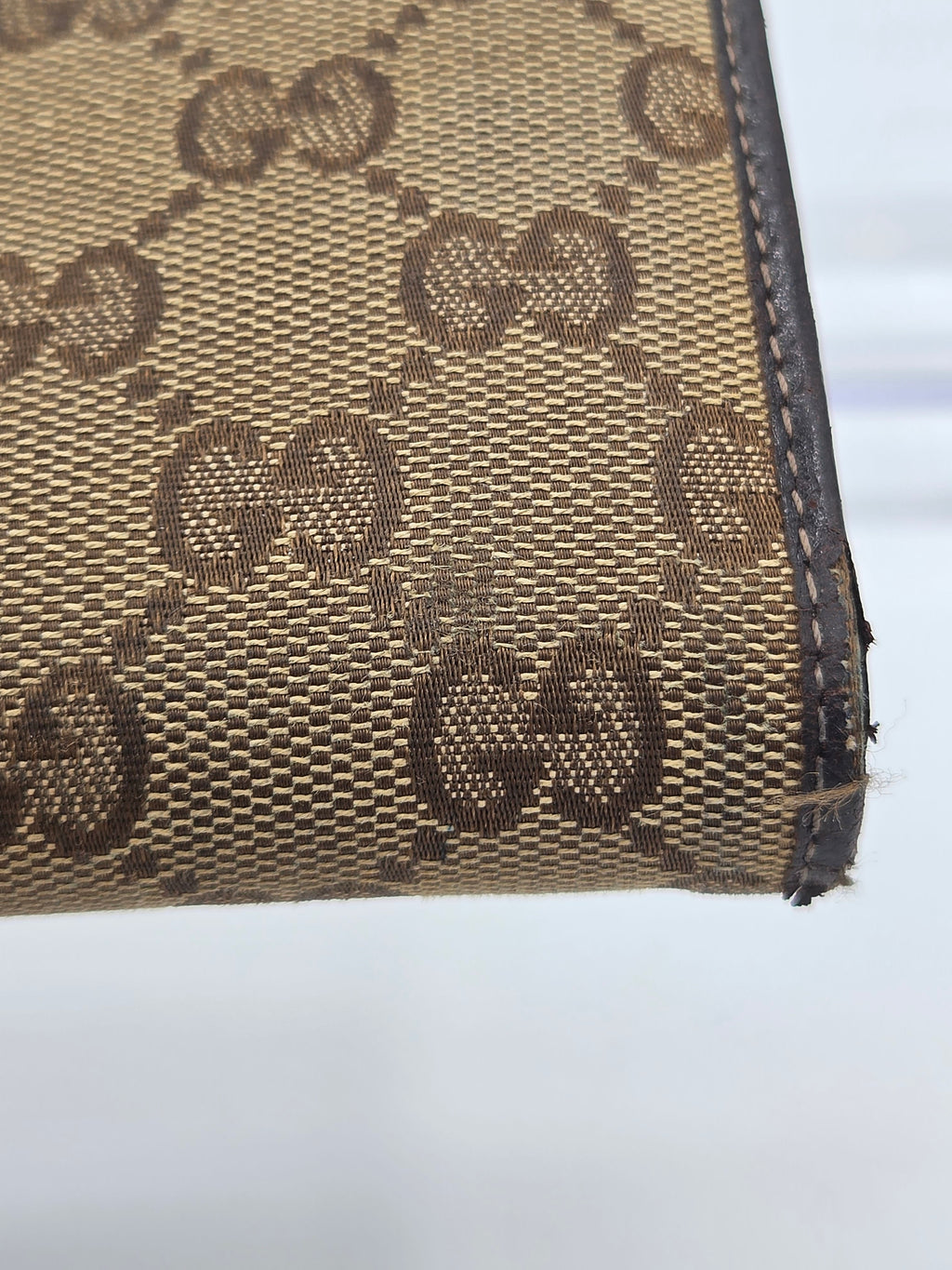Close-up of a beige and brown monogram canvas item with dark brown leather trim, showing the repeating interlocking G pattern and a frayed edge.