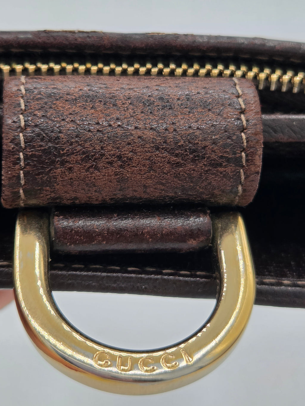 Close-up of a Gucci brown leather strap with a gold-tone metal D-ring and zipper pull, showing the Gucci logo engraved on the ring.