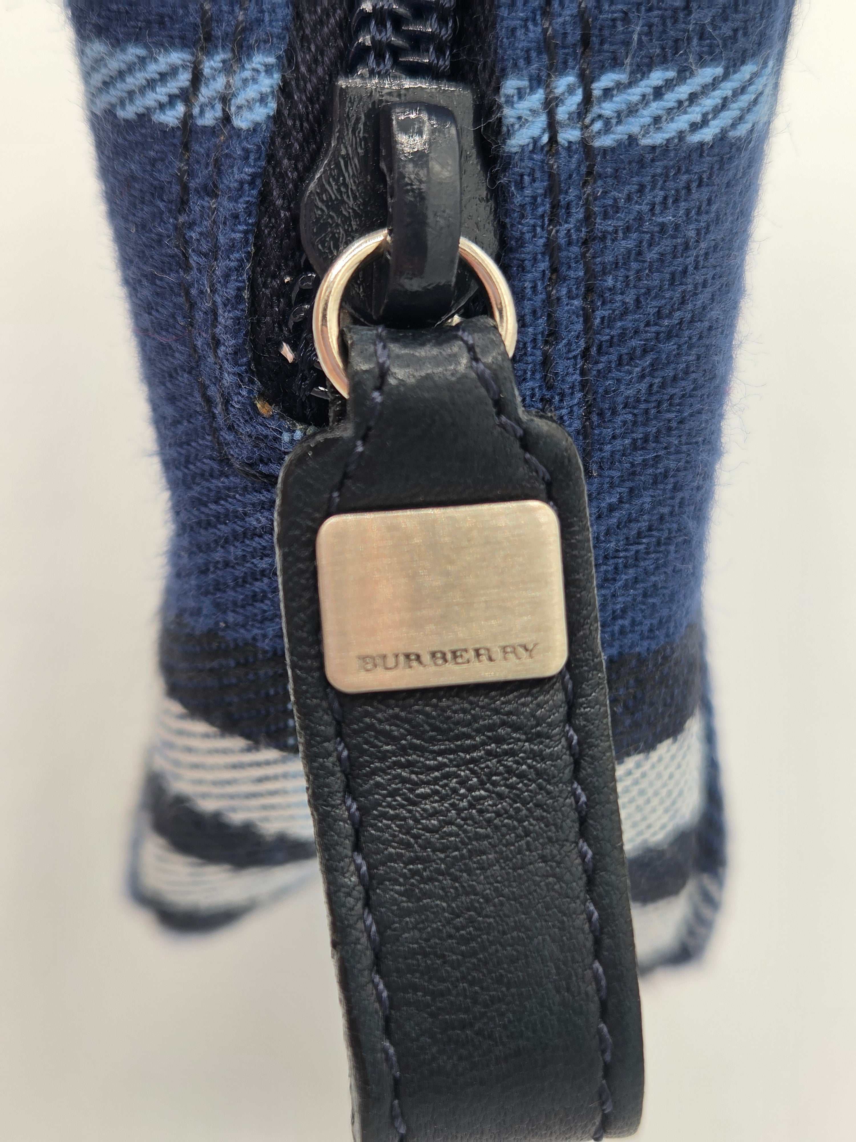 Close-up of a blue and white plaid fabric item with a black leather strap and a silver-tone metal plaque engraved with BURBERRY.