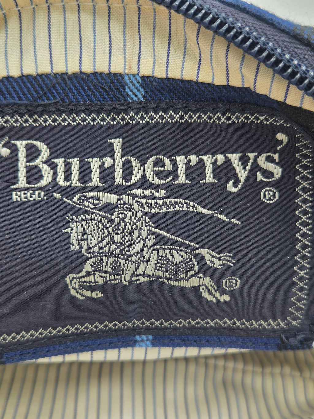 Close-up of a Burberry neck label sewn onto striped blue and beige fabric, showing the embroidered Burberry logo and text.