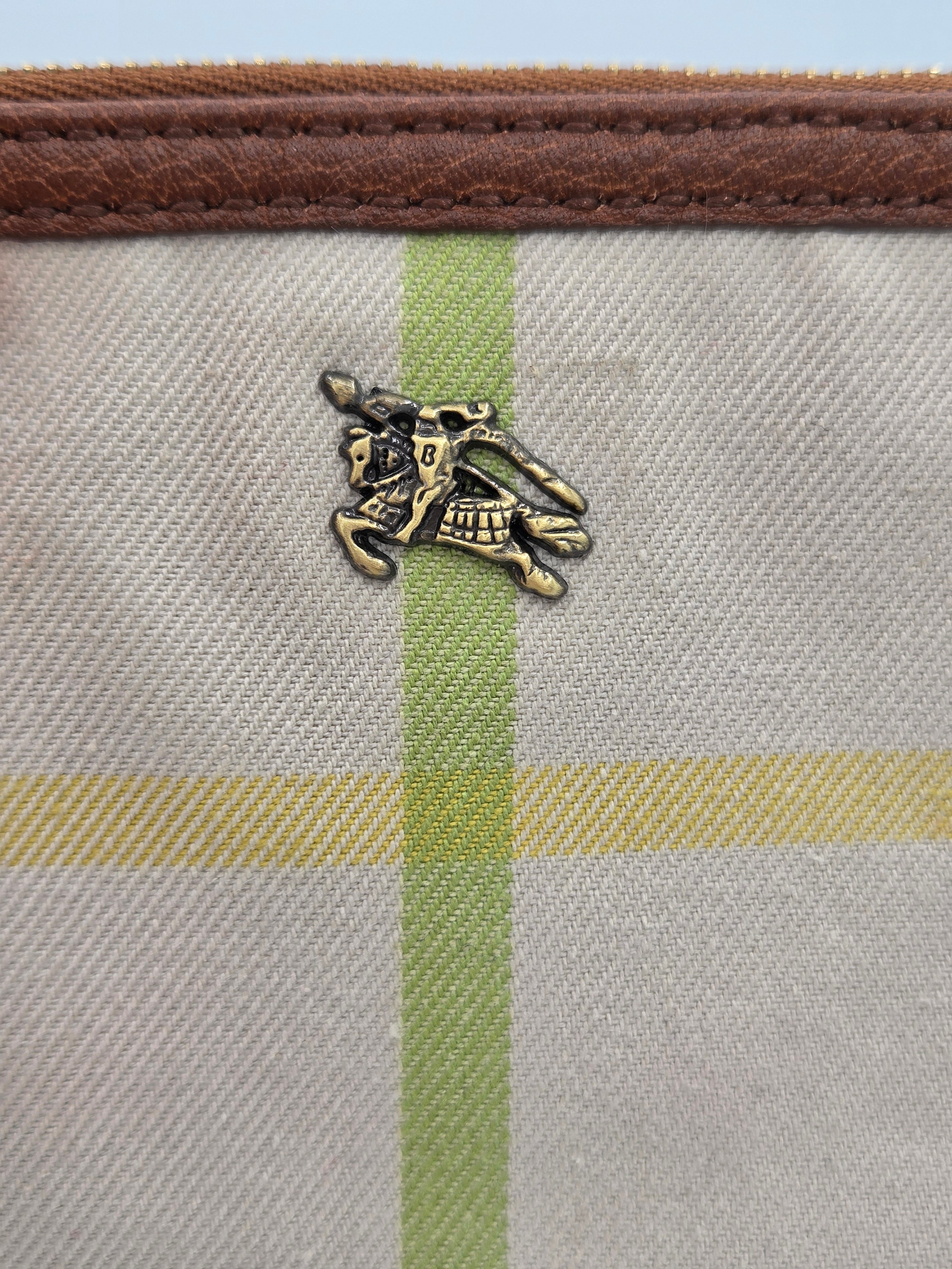 Close-up of a canvas pouch featuring a beige and green plaid pattern, a brown leather trim, and a small gold equestrian logo charm.