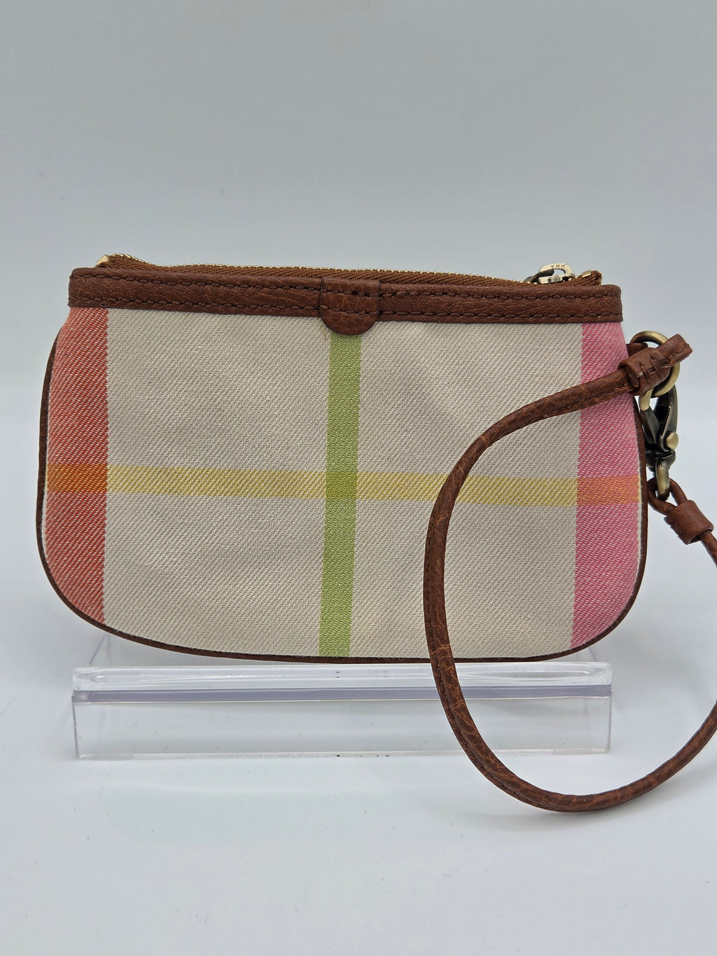A small plaid canvas wristlet clutch with brown leather trim and strap, featuring a pink, yellow, and green plaid pattern on a beige background, shown against a plain white wall.