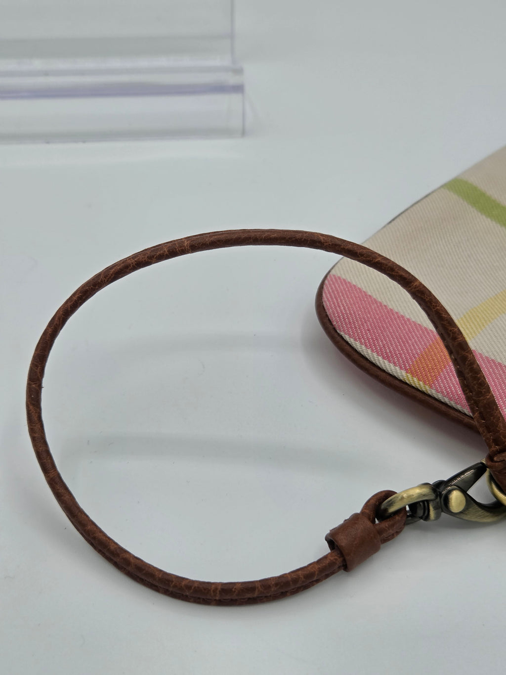 Close-up of a wristlet or clutch bag strap with a brown leather loop and a beige plaid pattern featuring pink, yellow, and green stripes, attached to a metal clasp.