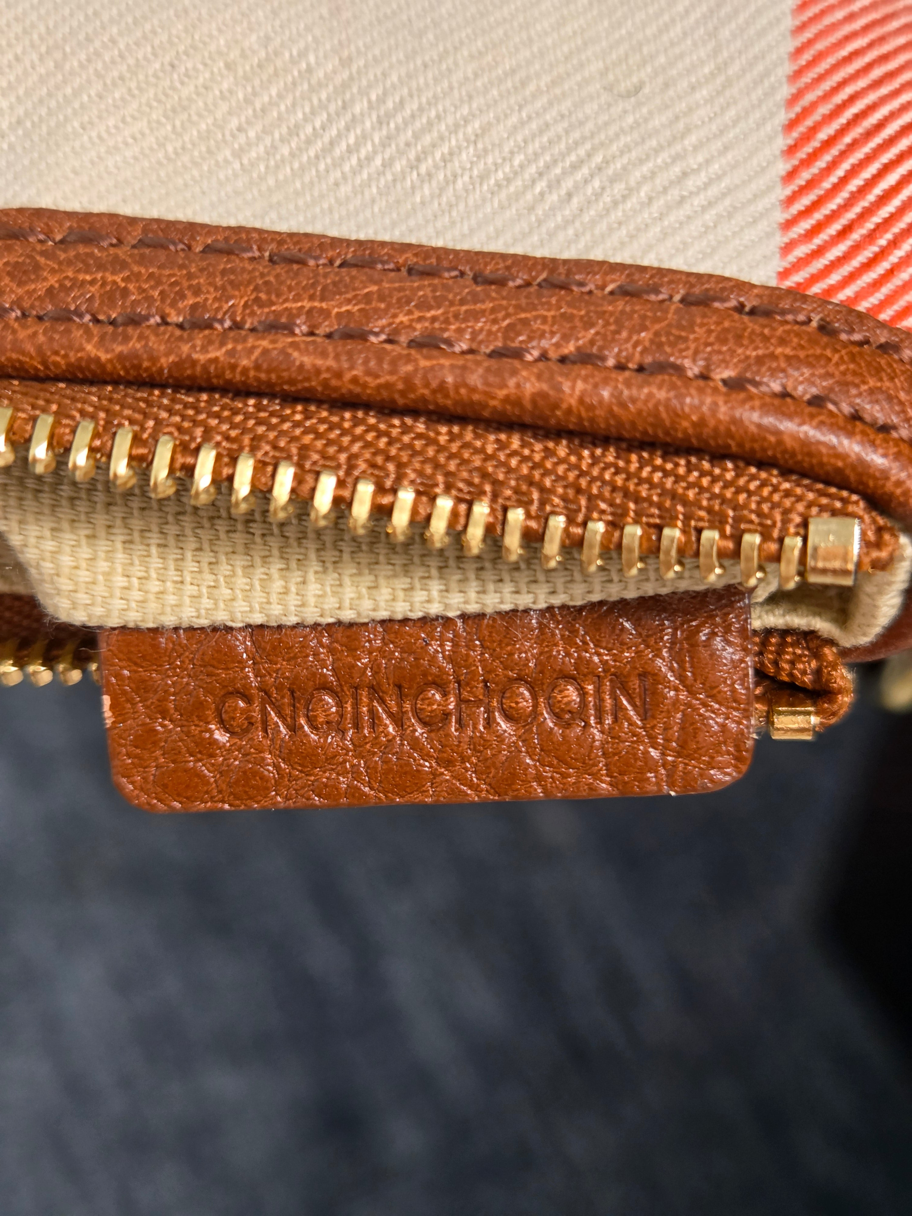 Close-up of a brown leather zipper pull tab with the brand name 'NICHOLAS JONES' embossed vertically, attached to a gold-tone zipper on a beige and red check patterned fabric.