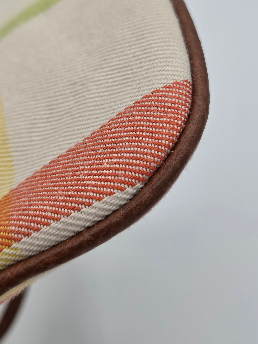 Close-up detail of a woven textile product showing a diagonal stripe pattern in terracotta, cream, and light yellow, finished with a smooth brown trim against a light gray background.