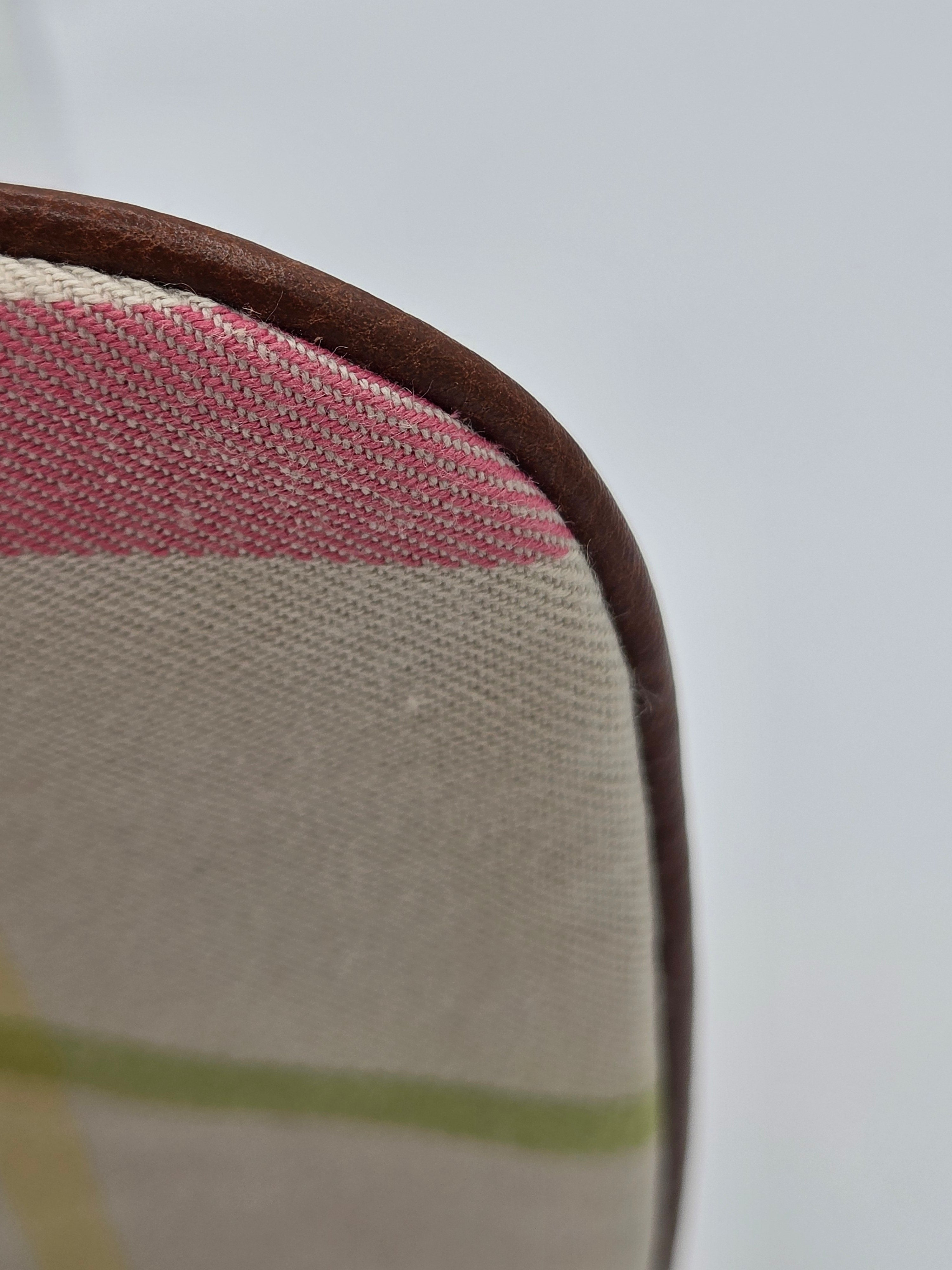 Close-up detail of a fabric cushion corner showing a beige woven textile with pink and light green stripes, finished with a dark brown stitched edge, against a plain light gray background.