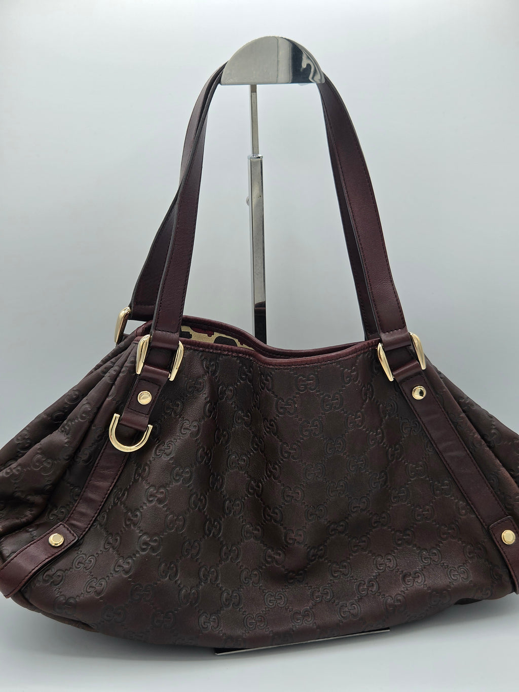 A dark brown monogram shoulder bag with dark brown leather straps and gold-tone hardware, displayed on a silver hanger against a light gray background.
