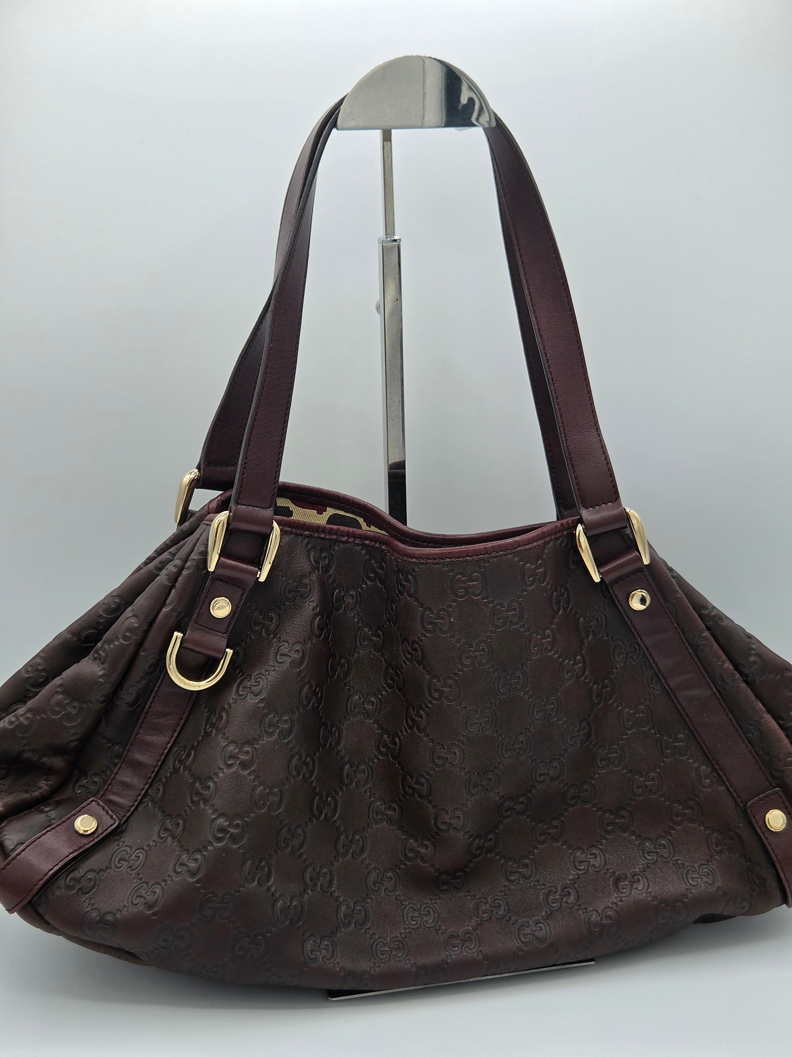 A dark brown monogram shoulder bag with dark brown leather straps and gold-tone hardware, displayed on a silver hanger against a light gray background.