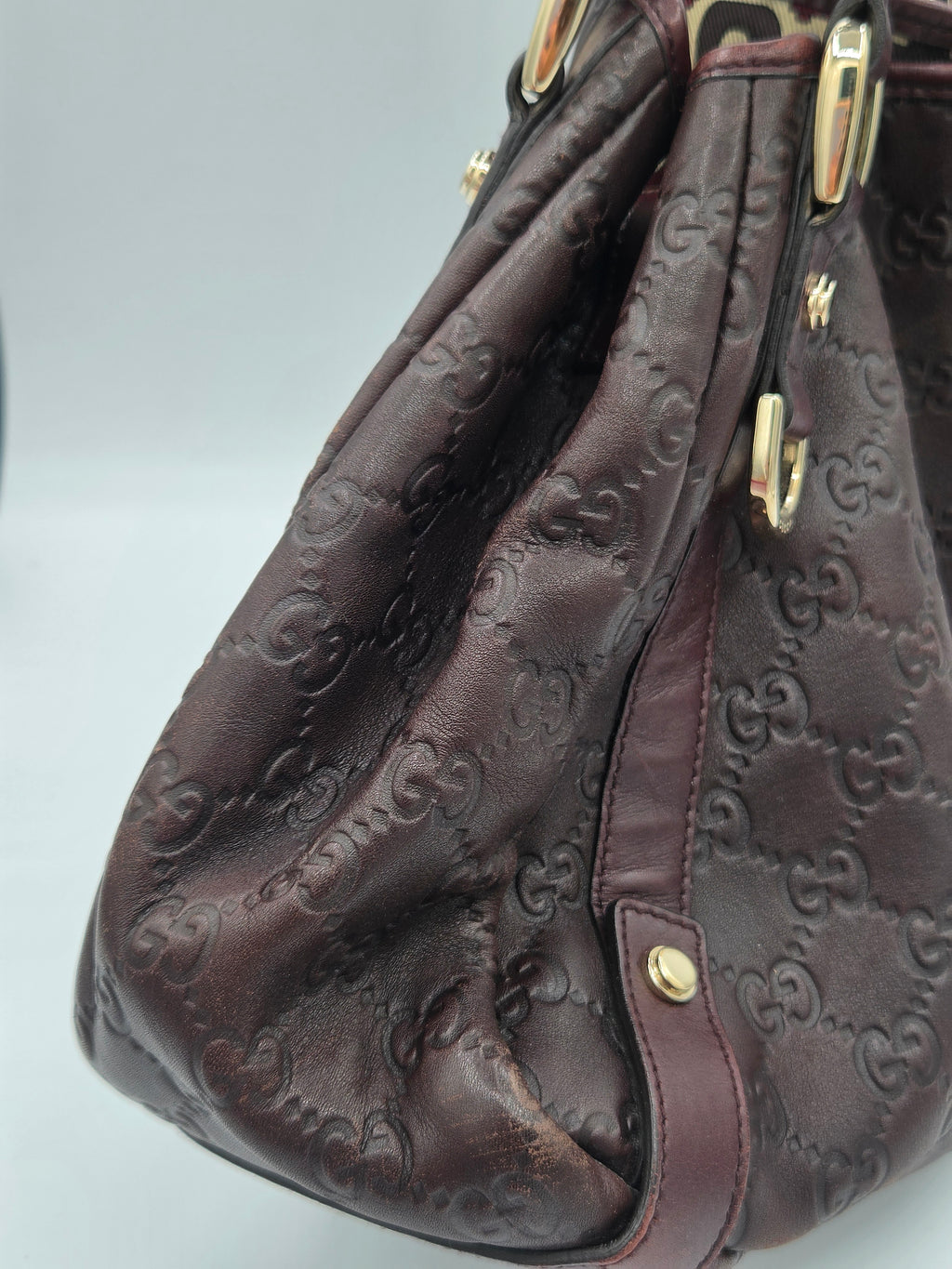 A close-up of a dark brown embossed leather handbag with gold-tone hardware and a repeating monogram pattern, set against a light gray background.