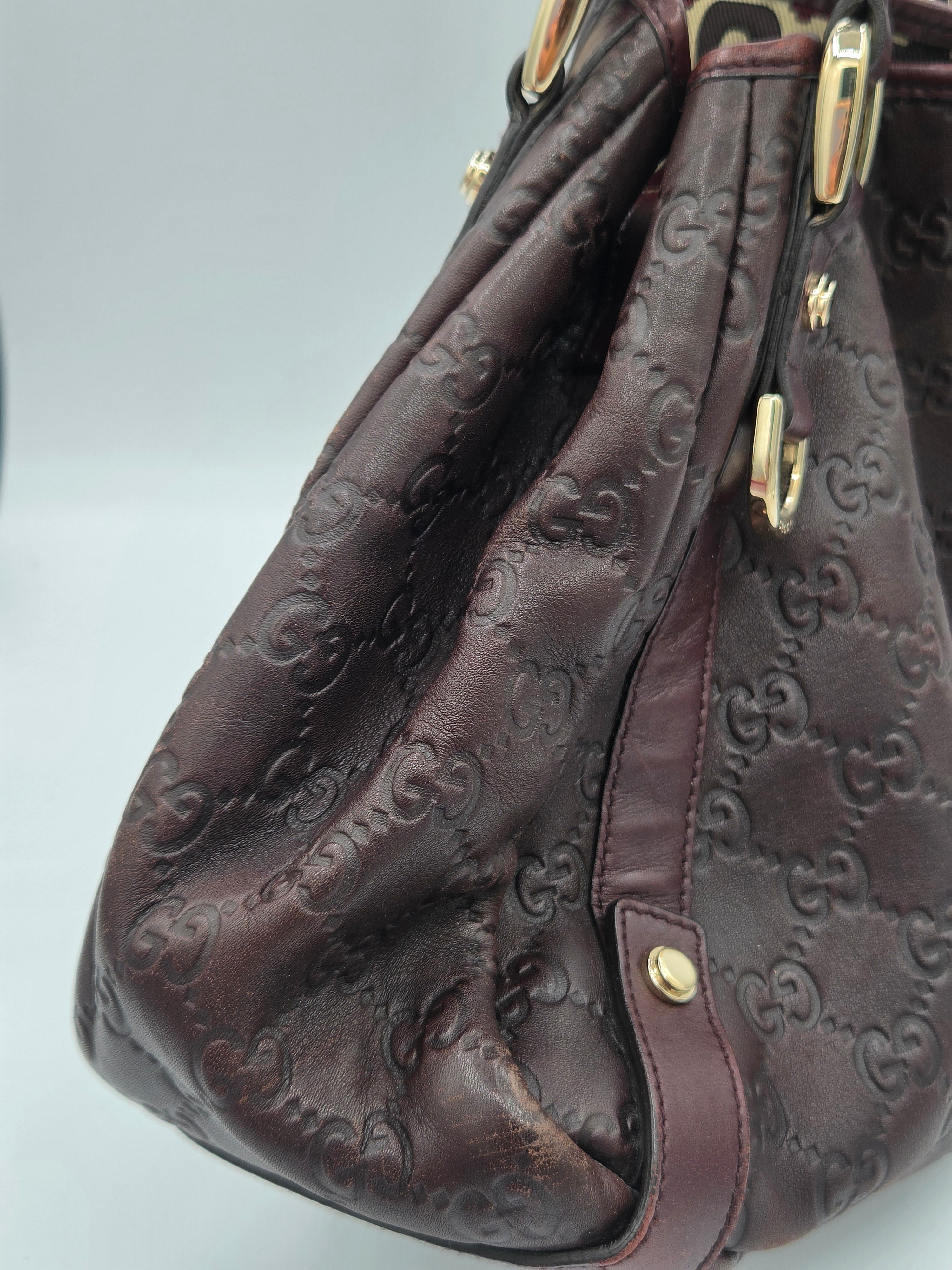A close-up of a dark brown embossed leather handbag with gold-tone hardware and a repeating monogram pattern, set against a light gray background.