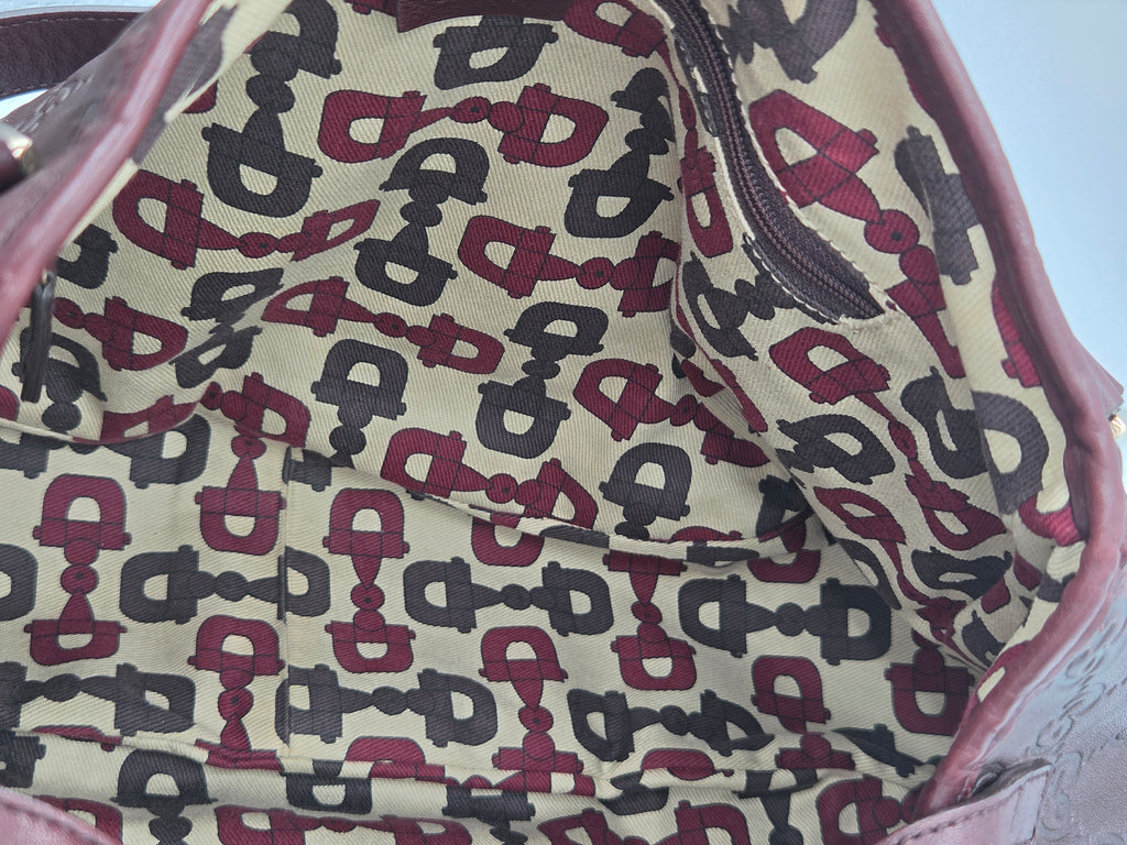 Close-up of the interior lining of a bag or pouch, featuring a repeating horse-bit pattern in burgundy and dark gray on a beige background, with a mauve-pink leather trim.