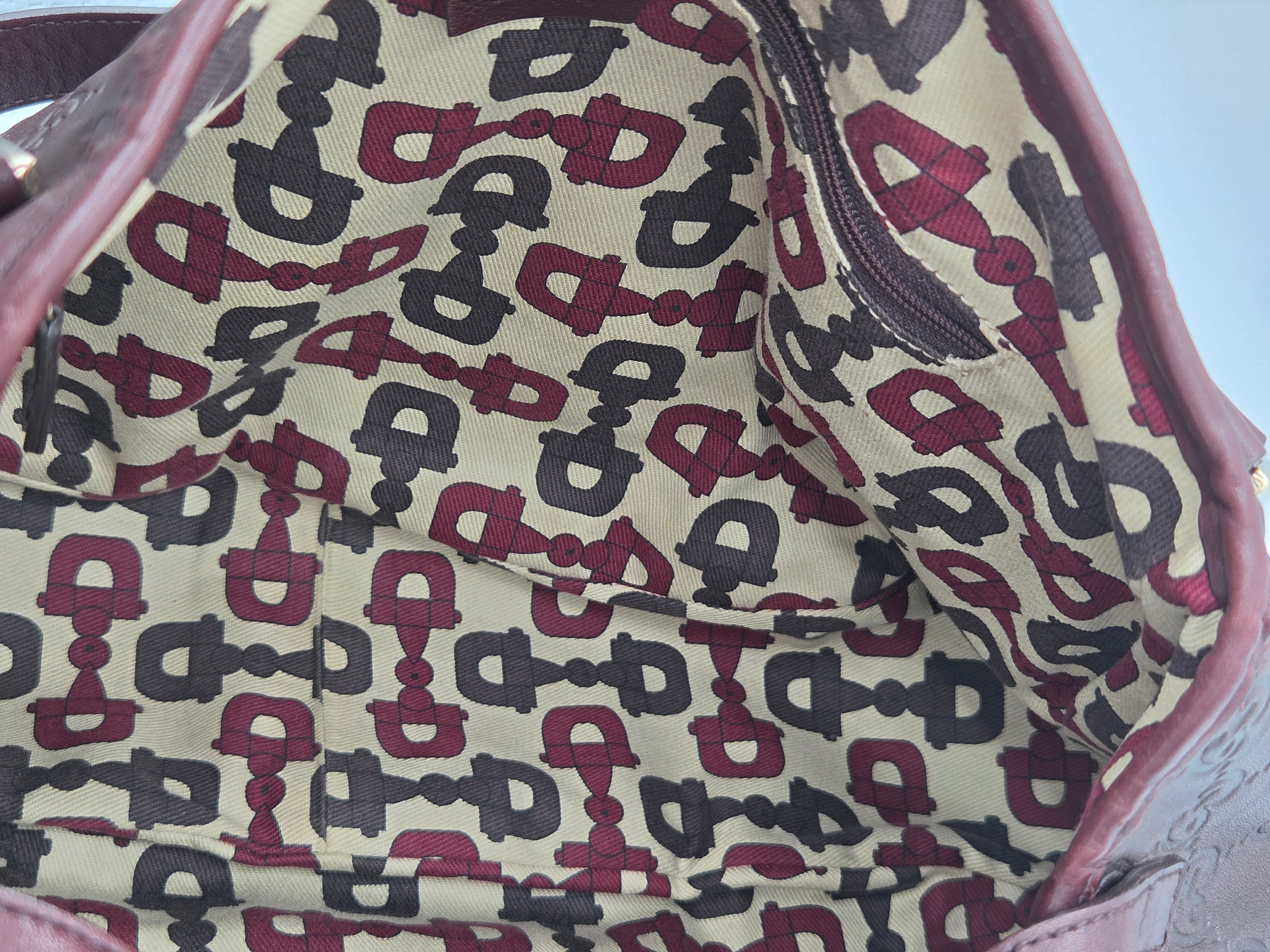 Close-up of the interior lining of a bag or pouch, featuring a repeating horse-bit pattern in burgundy and dark gray on a beige background, with a mauve-pink leather trim.