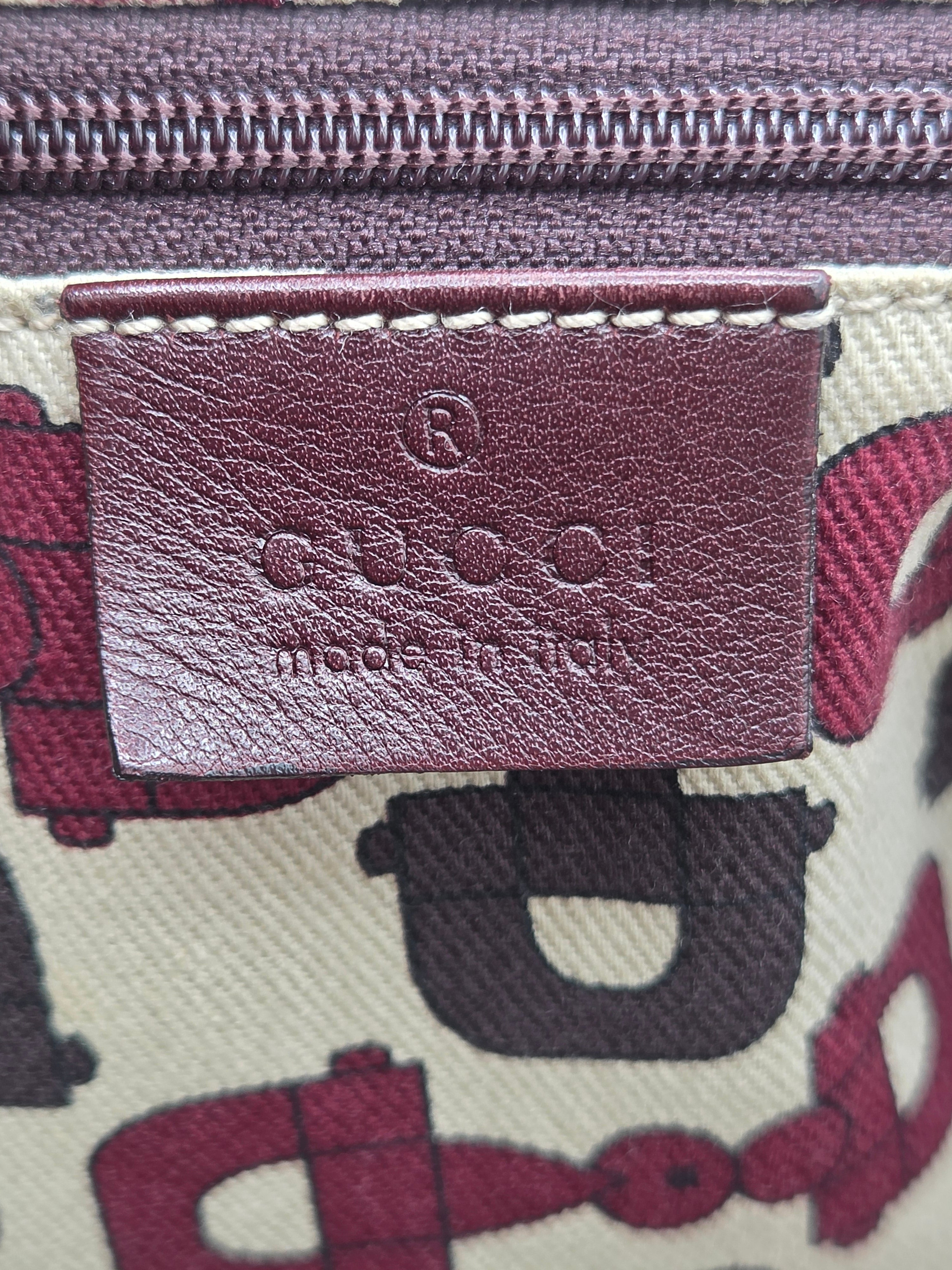 Close-up of a Coach zippered pouch or bag, showing a burgundy leather brand tab with embossed 'COACH made in USA' and a cream canvas body with a burgundy and black horse-and-carriage print.