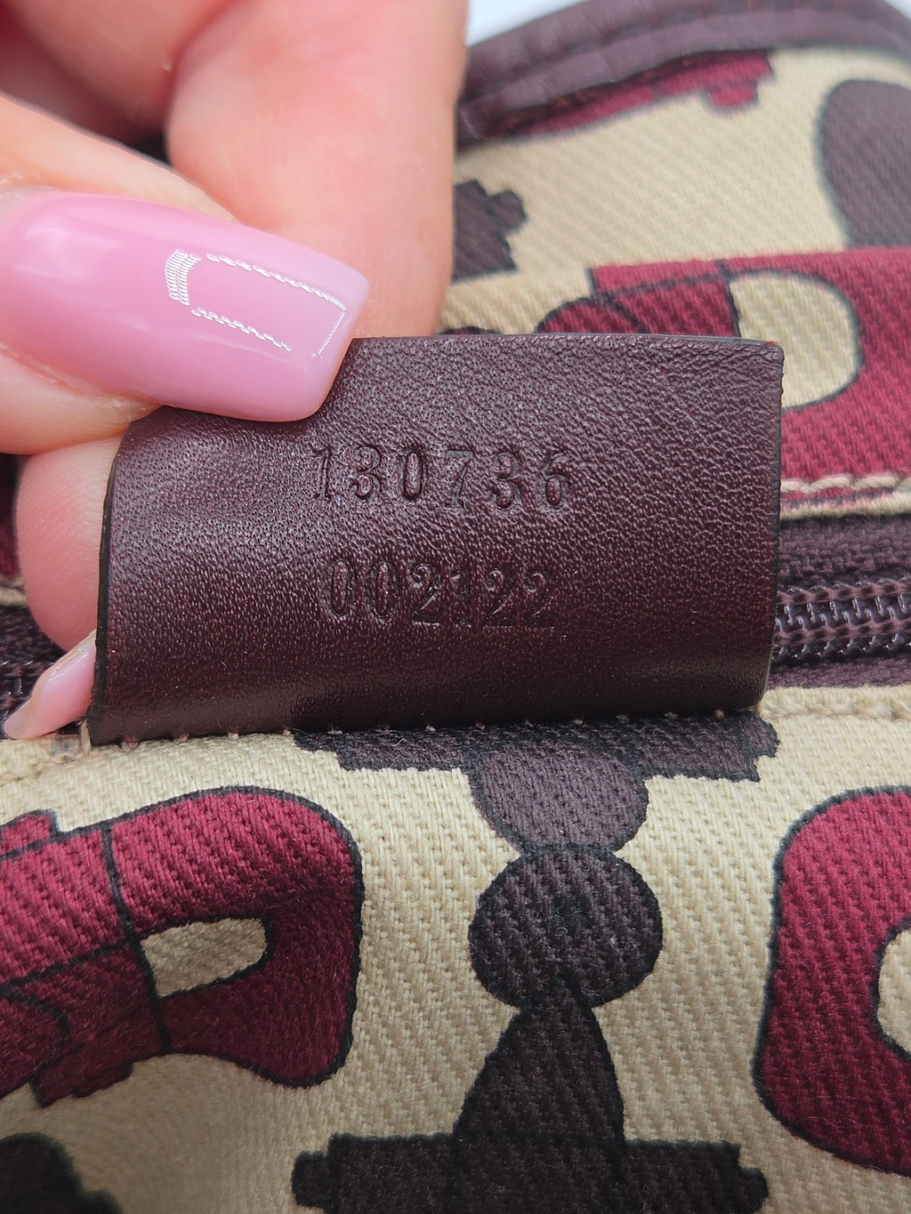 Close-up of a hand with pink manicured nails holding a brown leather zipper pull tab stamped with the number '130736 002122' over a beige canvas bag with a burgundy and black monogram pattern.