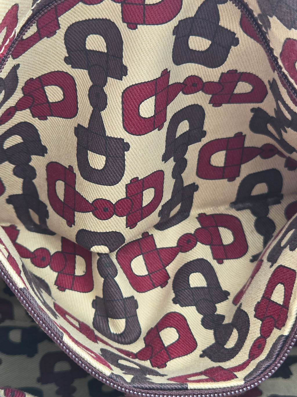 Close-up of a zipper pouch interior featuring an allover repeating horsebit pattern in burgundy and dark gray on a cream canvas background.