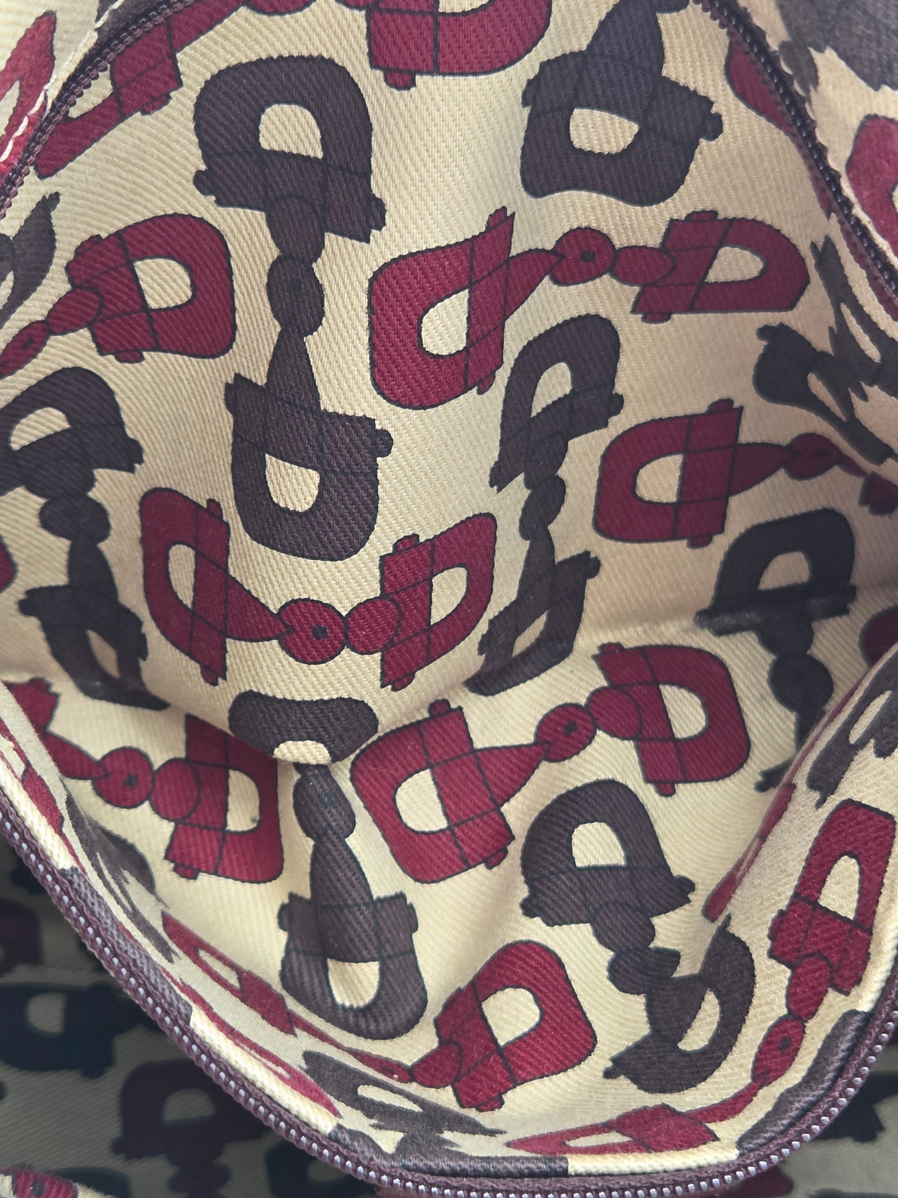Close-up of a zipper pouch interior featuring an allover repeating horsebit pattern in burgundy and dark gray on a cream canvas background.