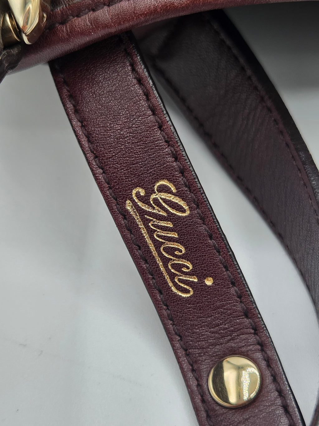 Close-up of a burgundy leather strap with gold-tone hardware and a gold Gucci logo embossed on the strap, set against a light gray background.
