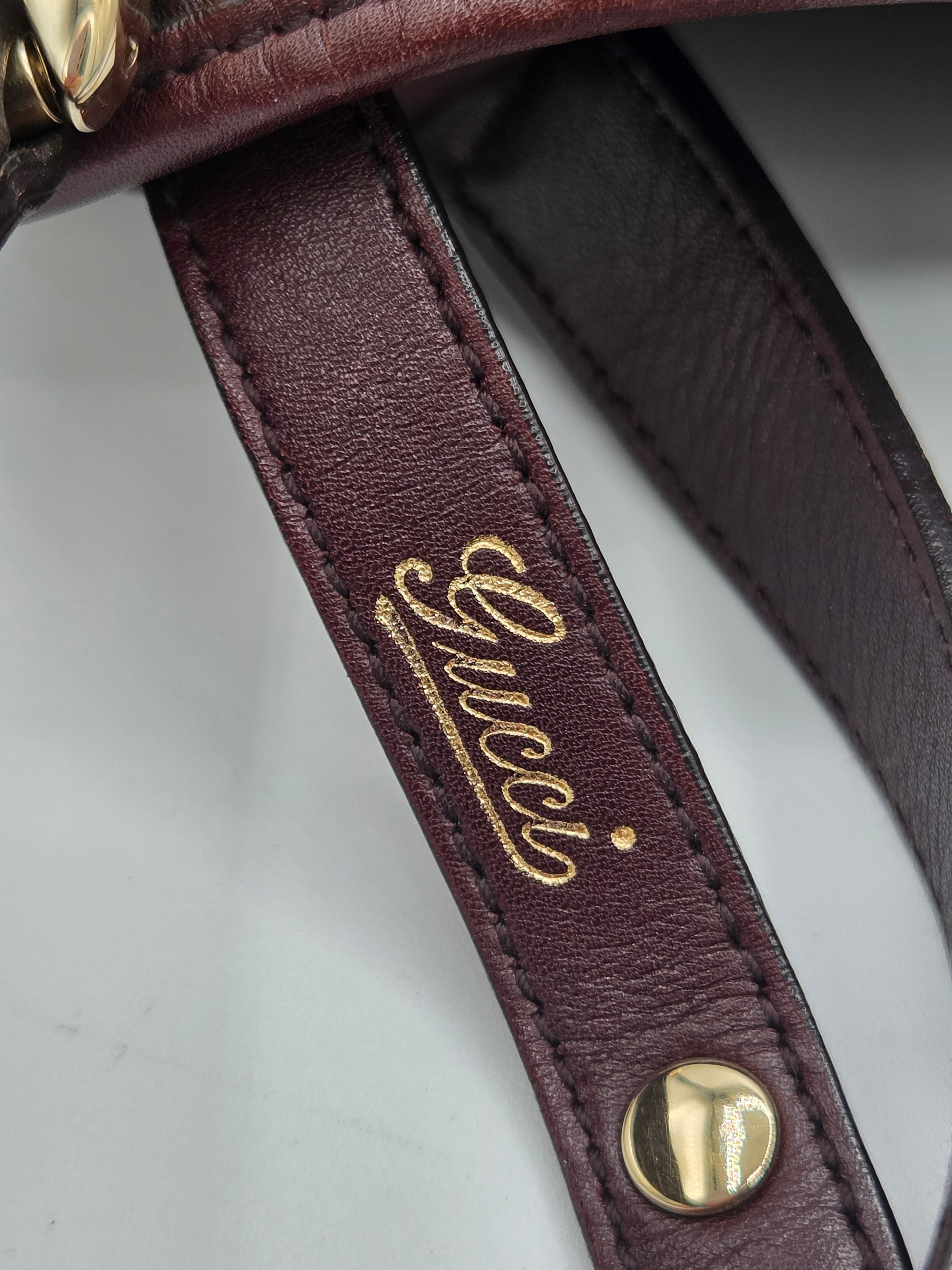 Close-up of a burgundy leather strap with gold-tone hardware and a gold Gucci logo embossed on the strap, set against a light gray background.