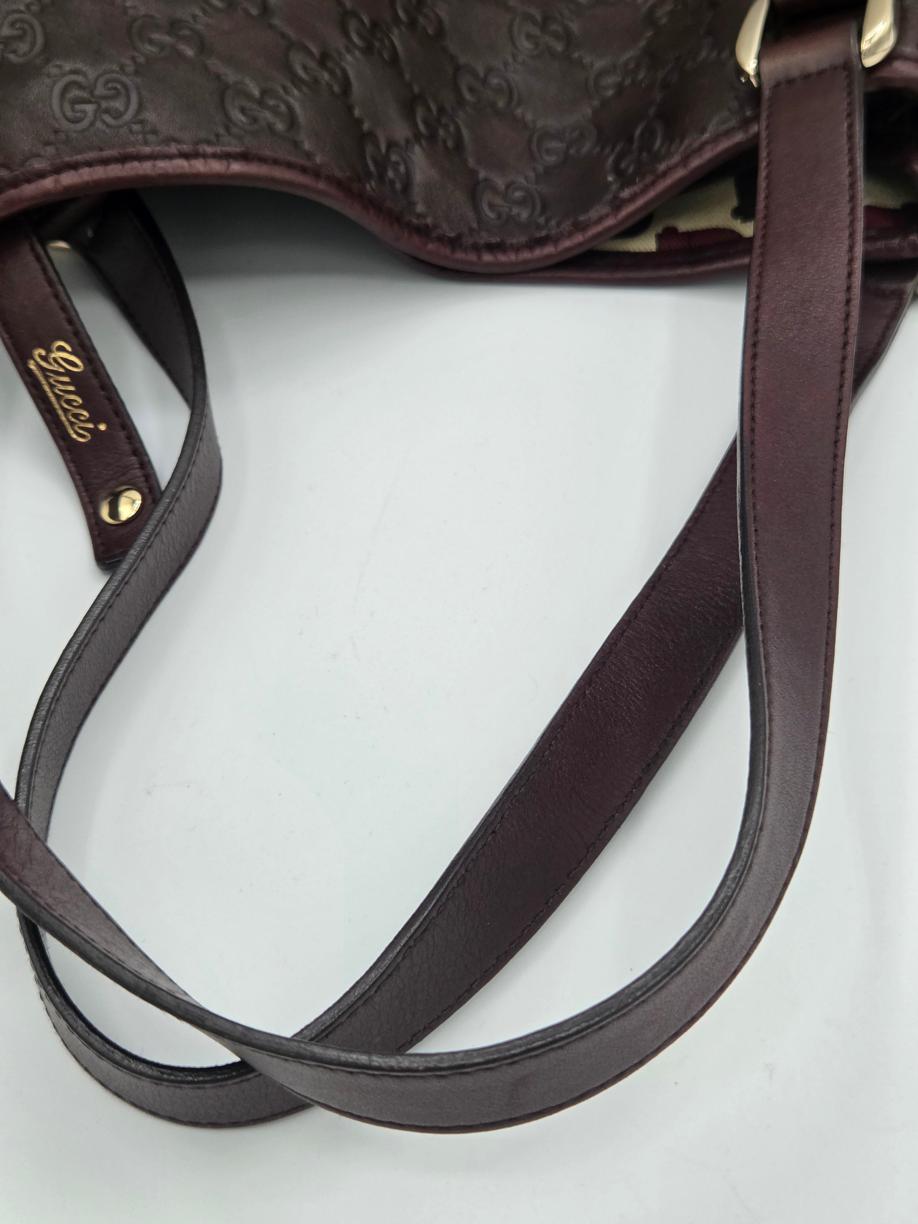 Close-up of a dark brown Gucci shoulder bag with embossed monogram pattern, a matching leather strap, and a small gold-tone Gucci logo tag on a light gray background.