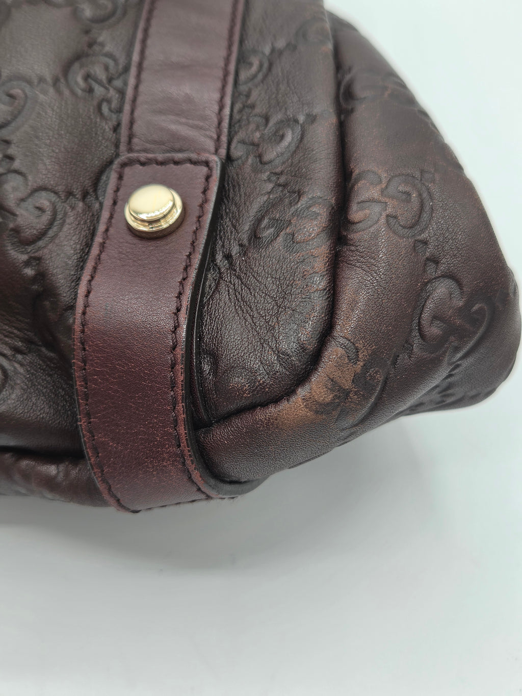 Close-up of a dark brown leather handbag with an embossed monogram pattern and a matching brown strap featuring a gold-tone snap button.