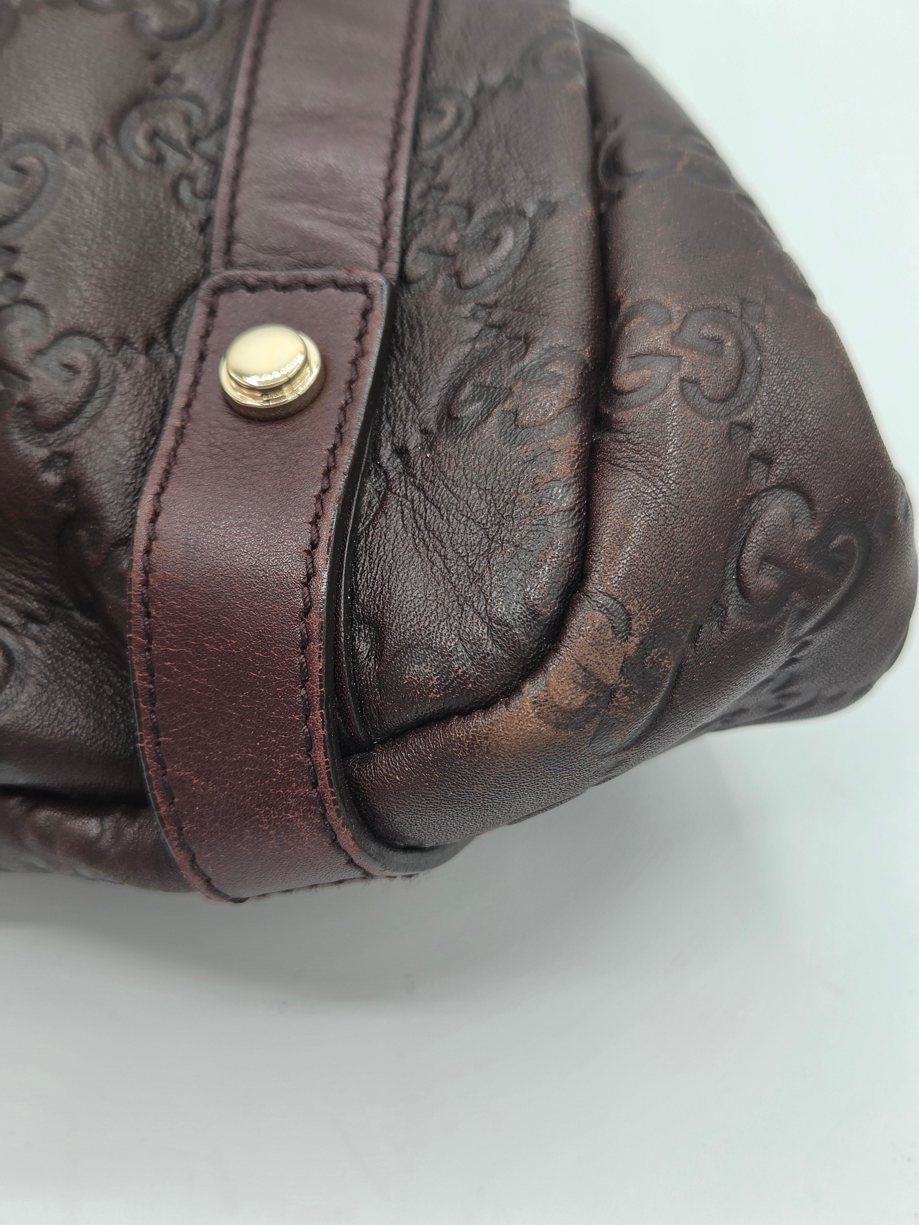 Close-up of a dark brown leather handbag with an embossed monogram pattern and a matching brown strap featuring a gold-tone snap button.