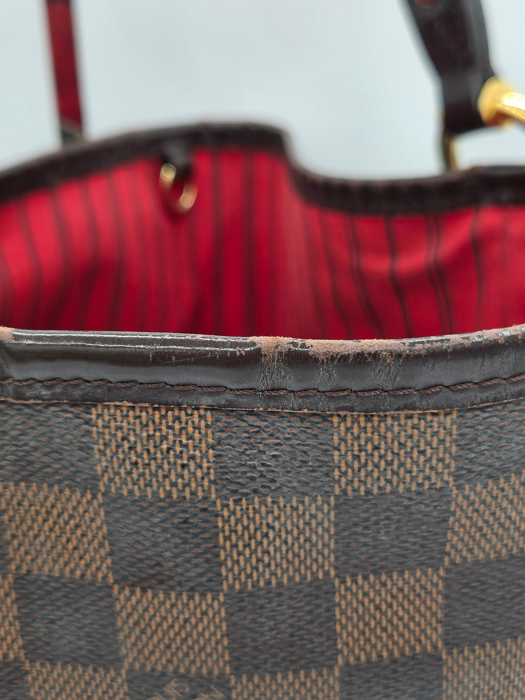 Close-up of a Damier Ebene checkered handbag showing the dark brown and beige canvas pattern, a dark brown leather trim, and a red striped fabric lining.