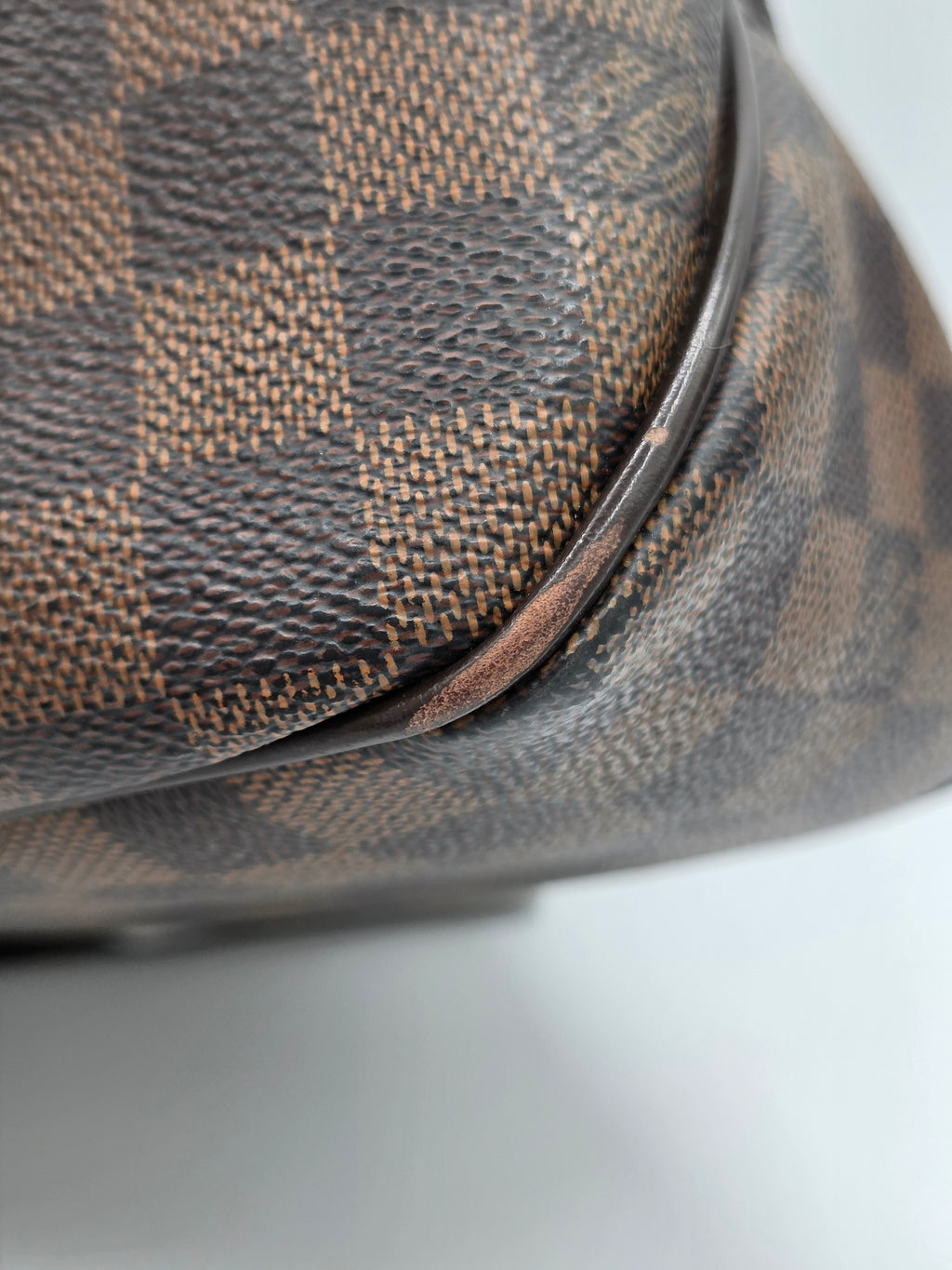 Close-up of a Louis Vuitton Damier canvas bag corner showing the classic brown and beige checkered pattern and dark brown leather trim.