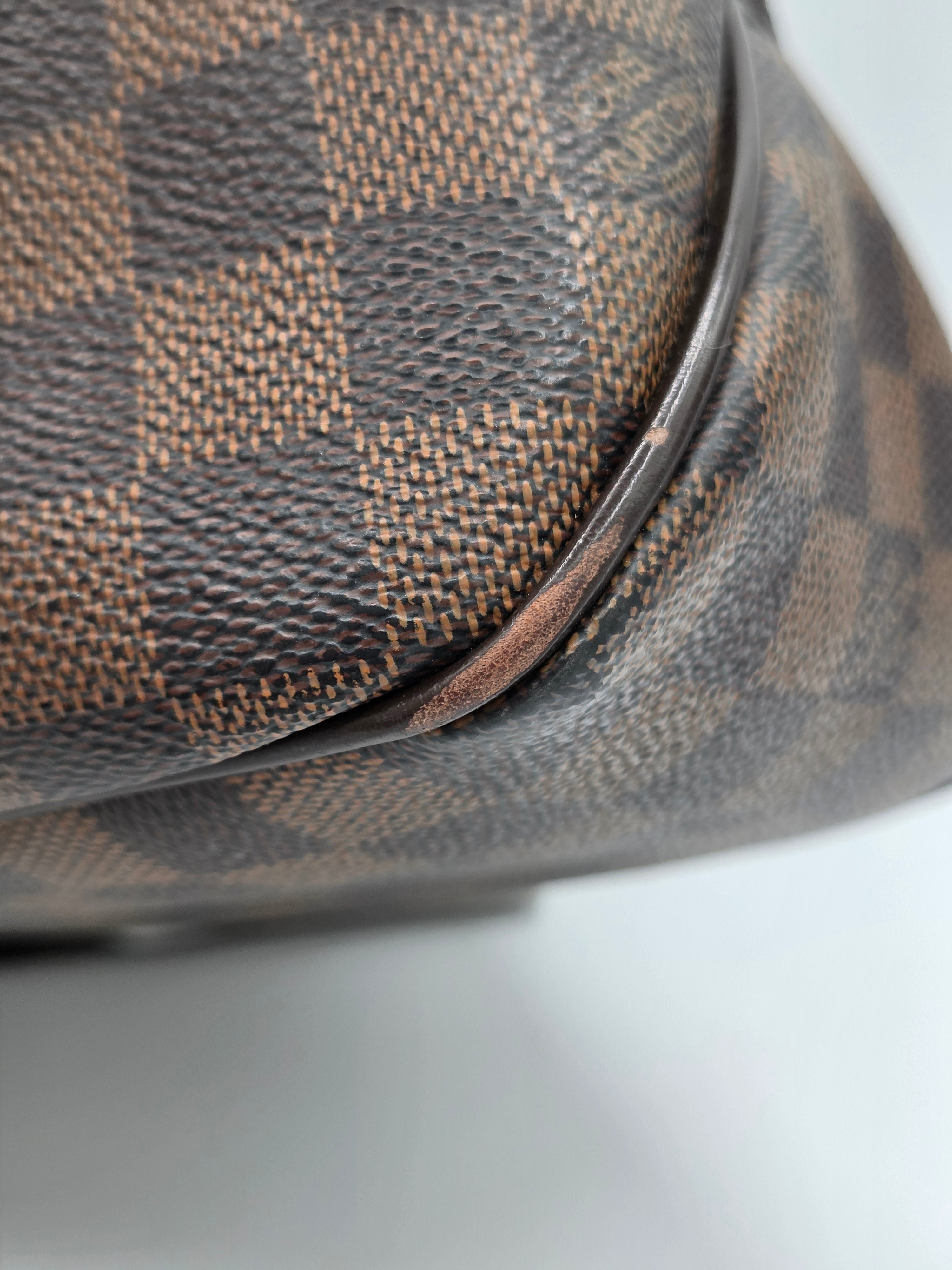 Close-up of a Louis Vuitton Damier canvas bag corner showing the classic brown and beige checkered pattern and dark brown leather trim.