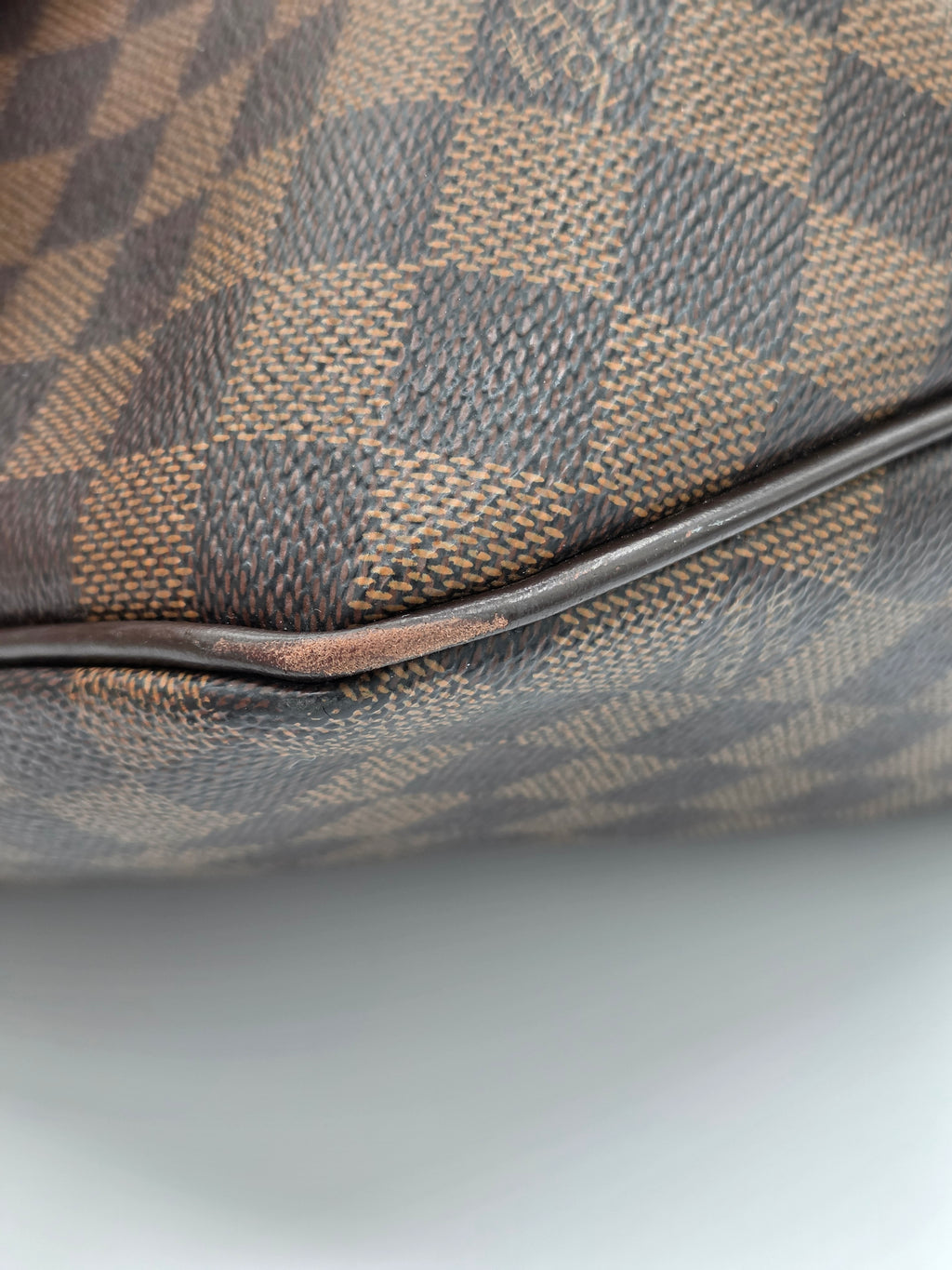 Close-up of a Louis Vuitton Damier canvas bag corner showing the classic brown and beige checkerboard pattern and a dark brown leather piping edge.