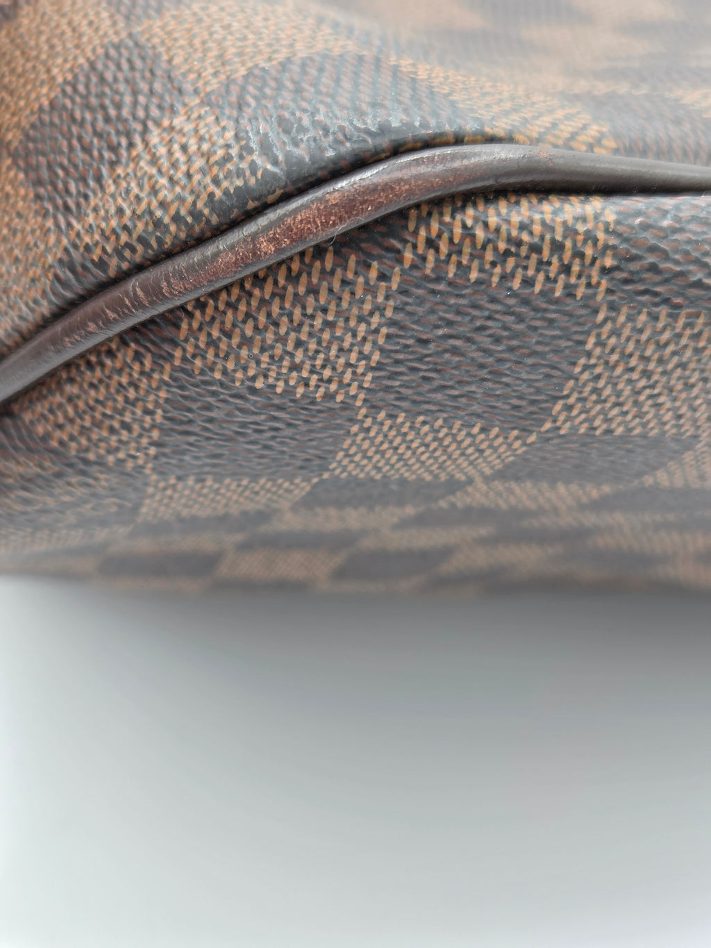 Close-up of a Louis Vuitton Damier canvas bag corner showing the classic brown and gray checkered pattern and a dark brown leather trim.