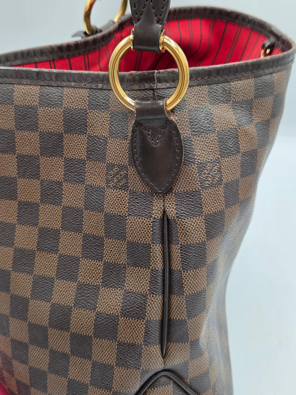 Close-up of a Louis Vuitton handbag in Damier Ebene canvas, showing the brown and beige checkered pattern, dark brown leather trim, a gold-tone ring detail, and a red striped interior lining.