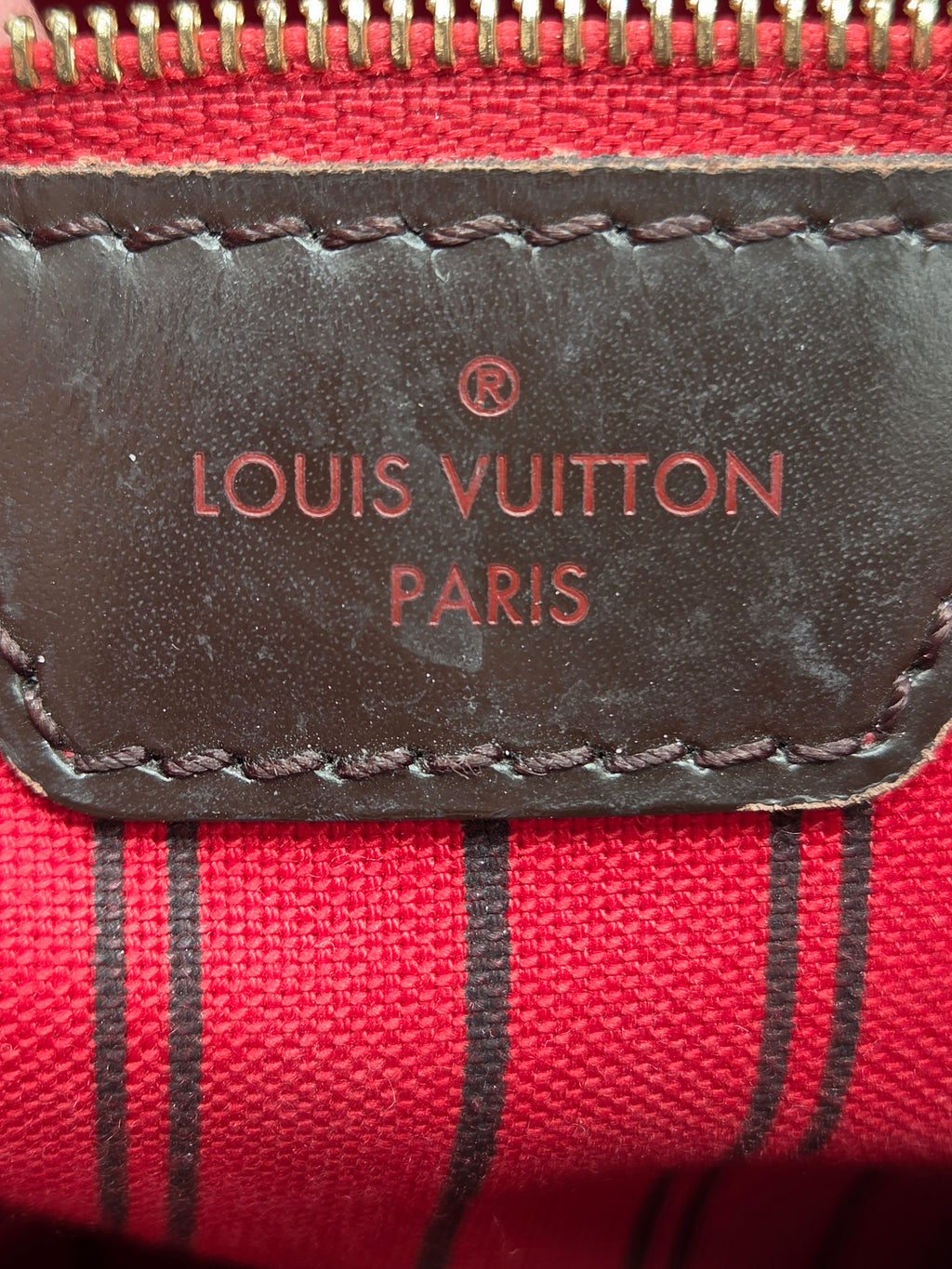 Close-up of a Louis Vuitton pouch showing a dark brown leather patch with embossed red 'LOUIS VUITTON PARIS' text, stitched onto a red canvas background with thin black vertical stripes.